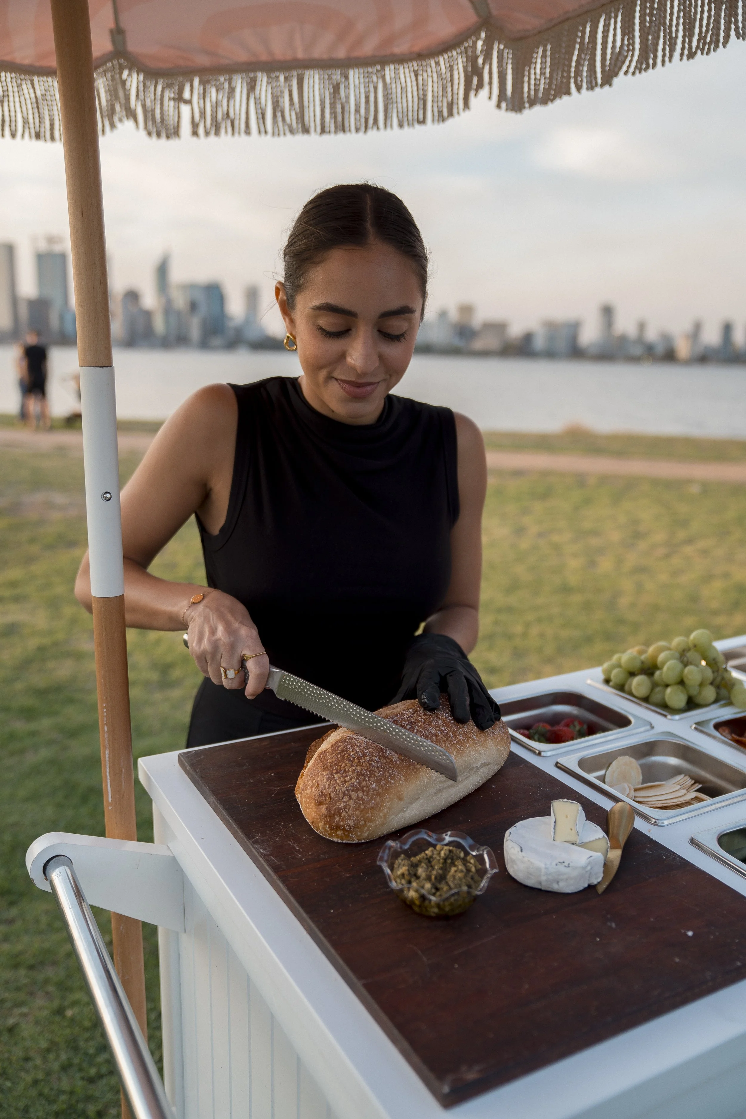 A woman with dark hair pulled back, wearing a black sleeveless top, is slicing a loaf of bread at an outdoor food stand under a pink umbrella. The stand has a wooden surface with various cheeses, grapes, and condiments. In the background, there is a 