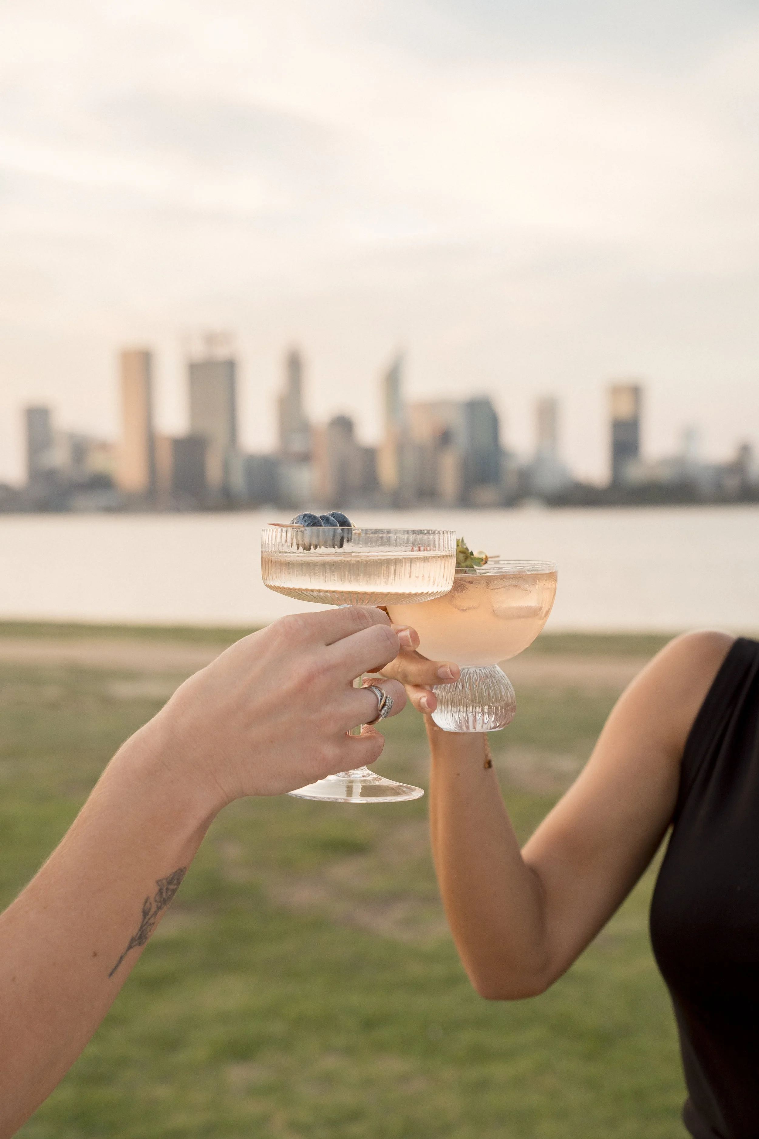 Two people clinking champagne glasses with blueberries and a sprig of mint inside, outdoors near a body of water, with a city skyline in the background during sunset.