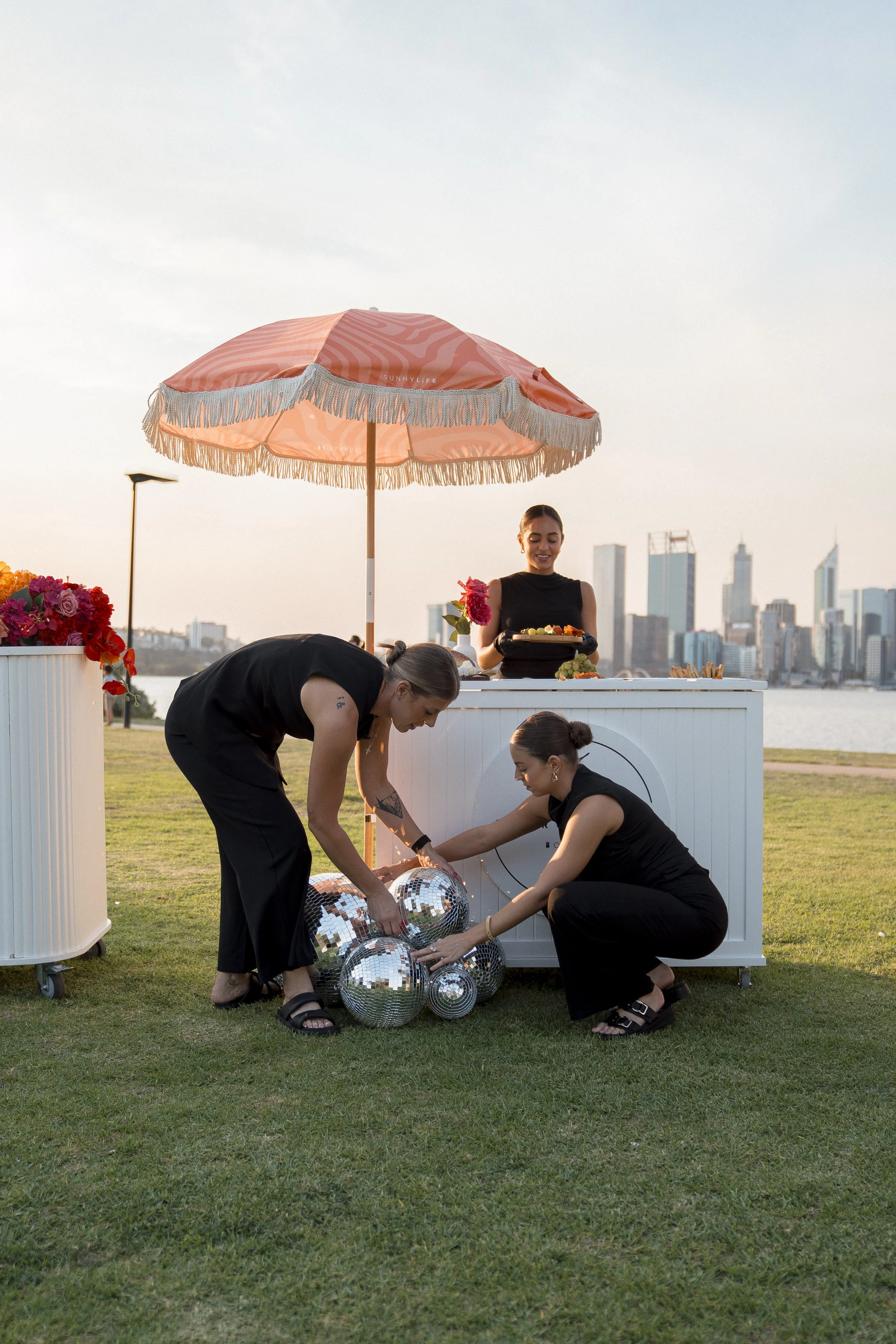 People setting up disco balls and decorations outdoors near a city skyline at sunset, with a pink parasol providing shade.