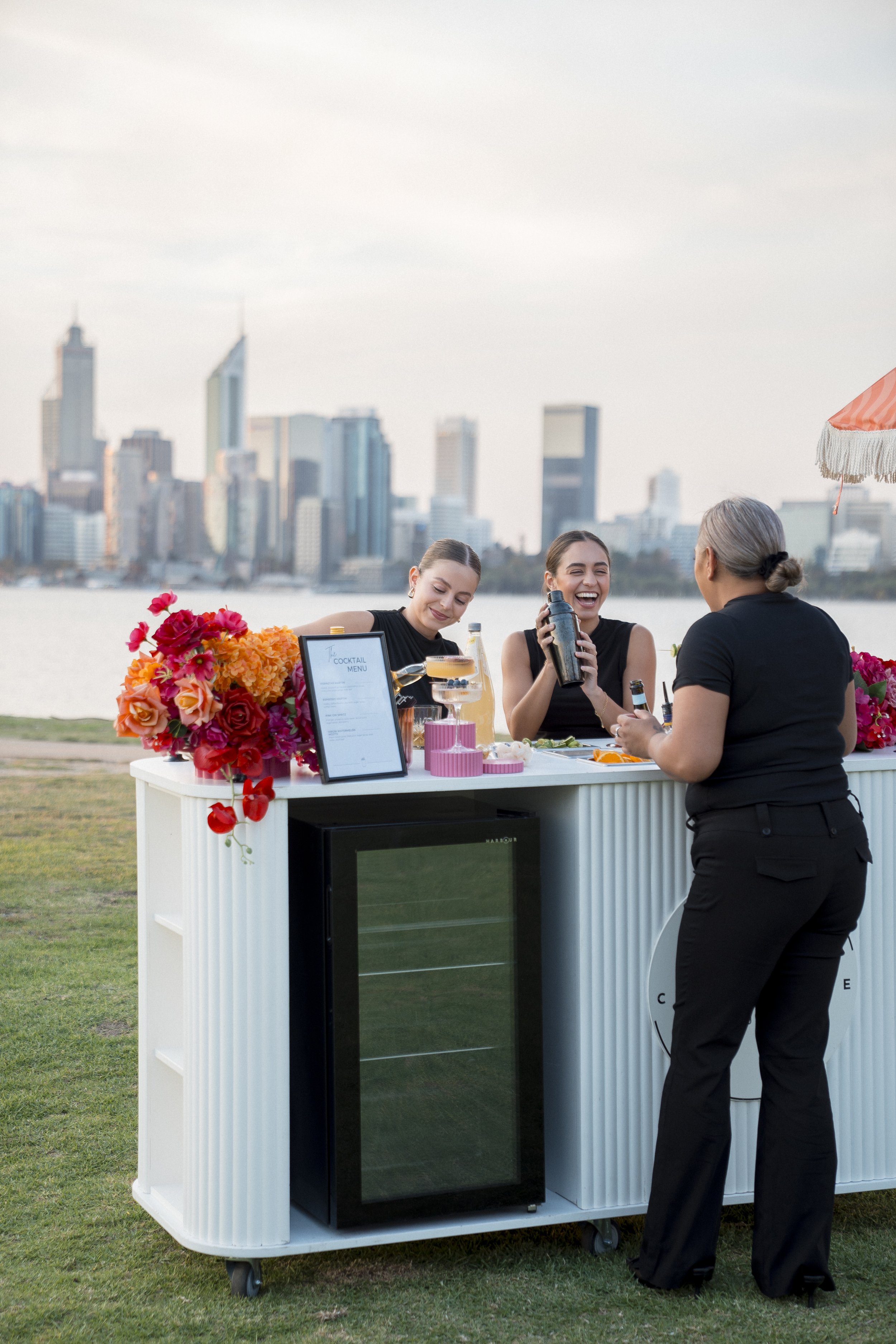Three women sitting at a drink station with a city skyline in the background. The station features a black drink dispenser and a floral decoration, and a young woman behind the station is serving drinks.