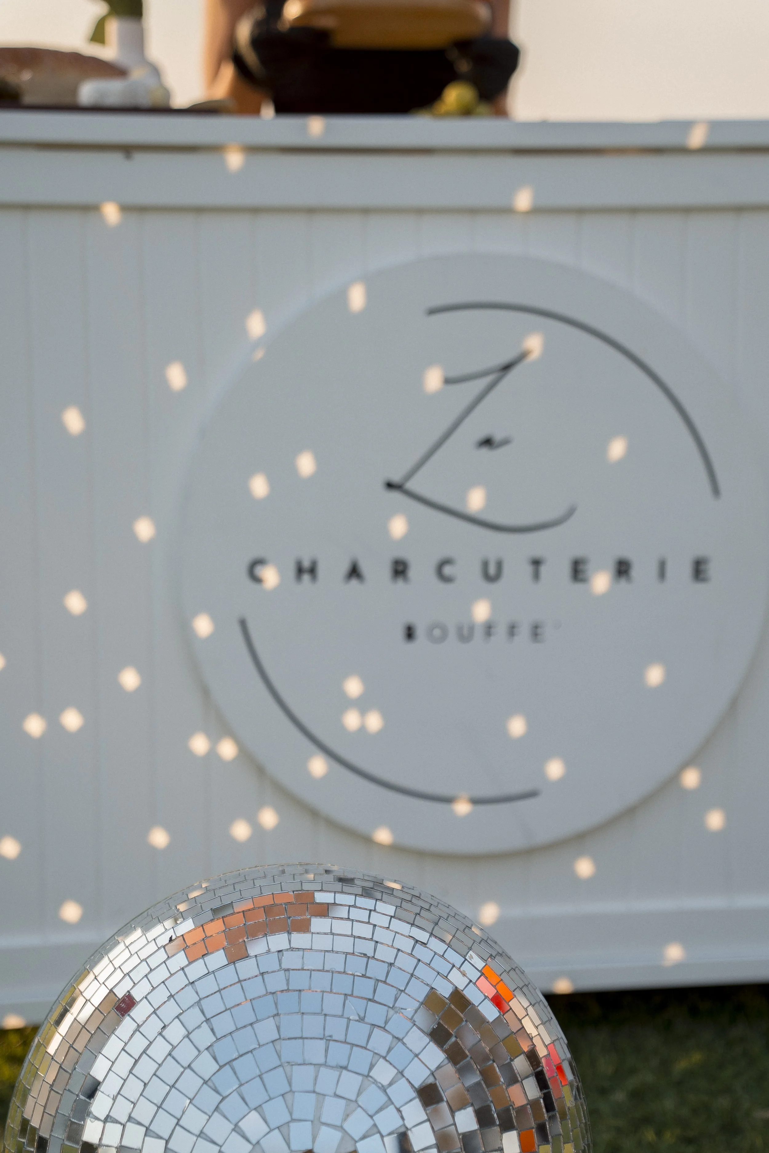 Close-up of a silver disco ball reflecting light, with a blurred background showing a decorative sign that reads 'Charcuterie Bouffe' and a wall with small, evenly spaced lights.