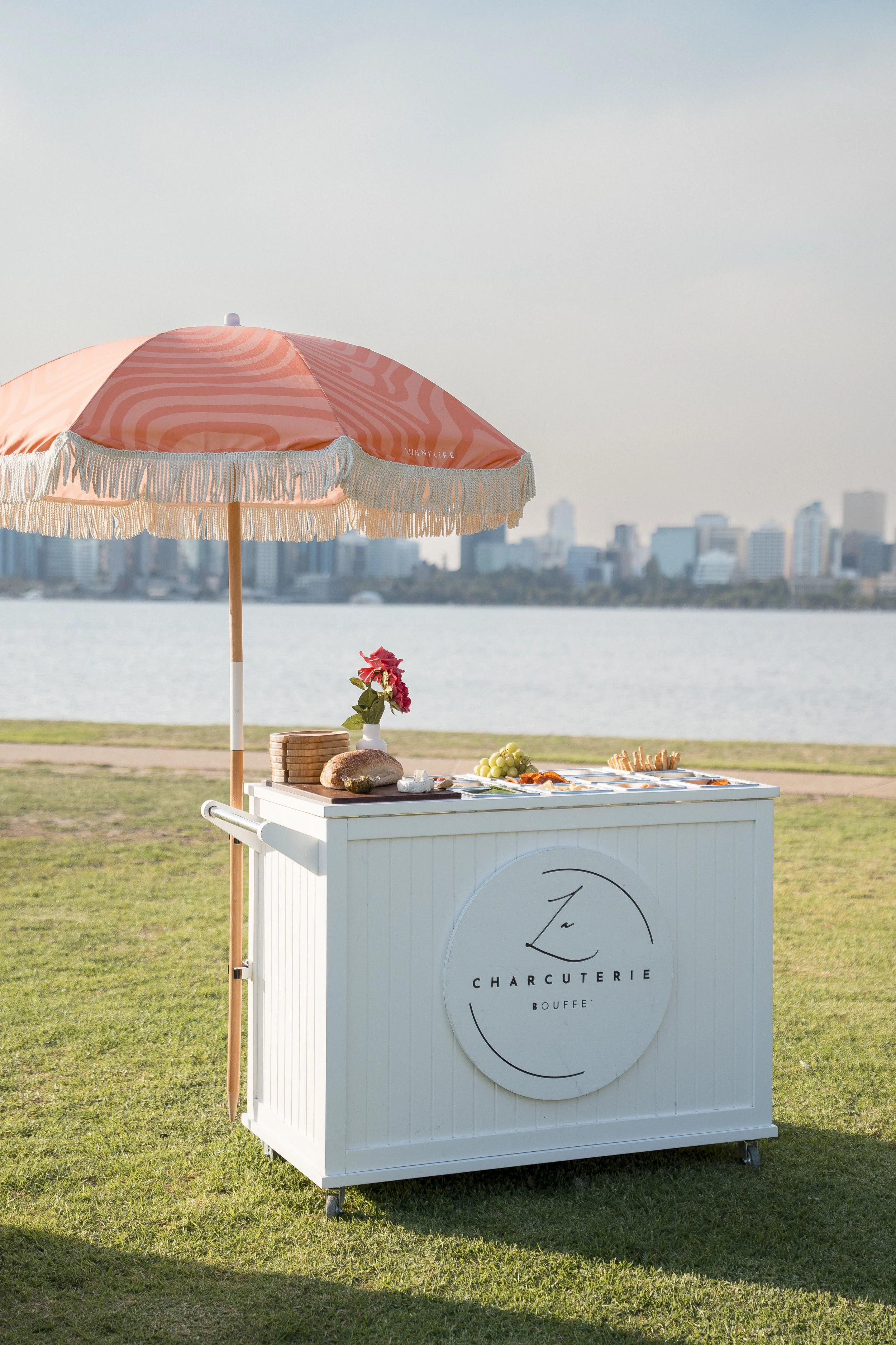 A white food cart with a circular logo that reads "Charcuterie Bouff" situated on a grassy area near a body of water. The cart has a pink and beige striped beach umbrella with fringe, a small vase with pink flowers, grapes, bread, cheese, and cracker