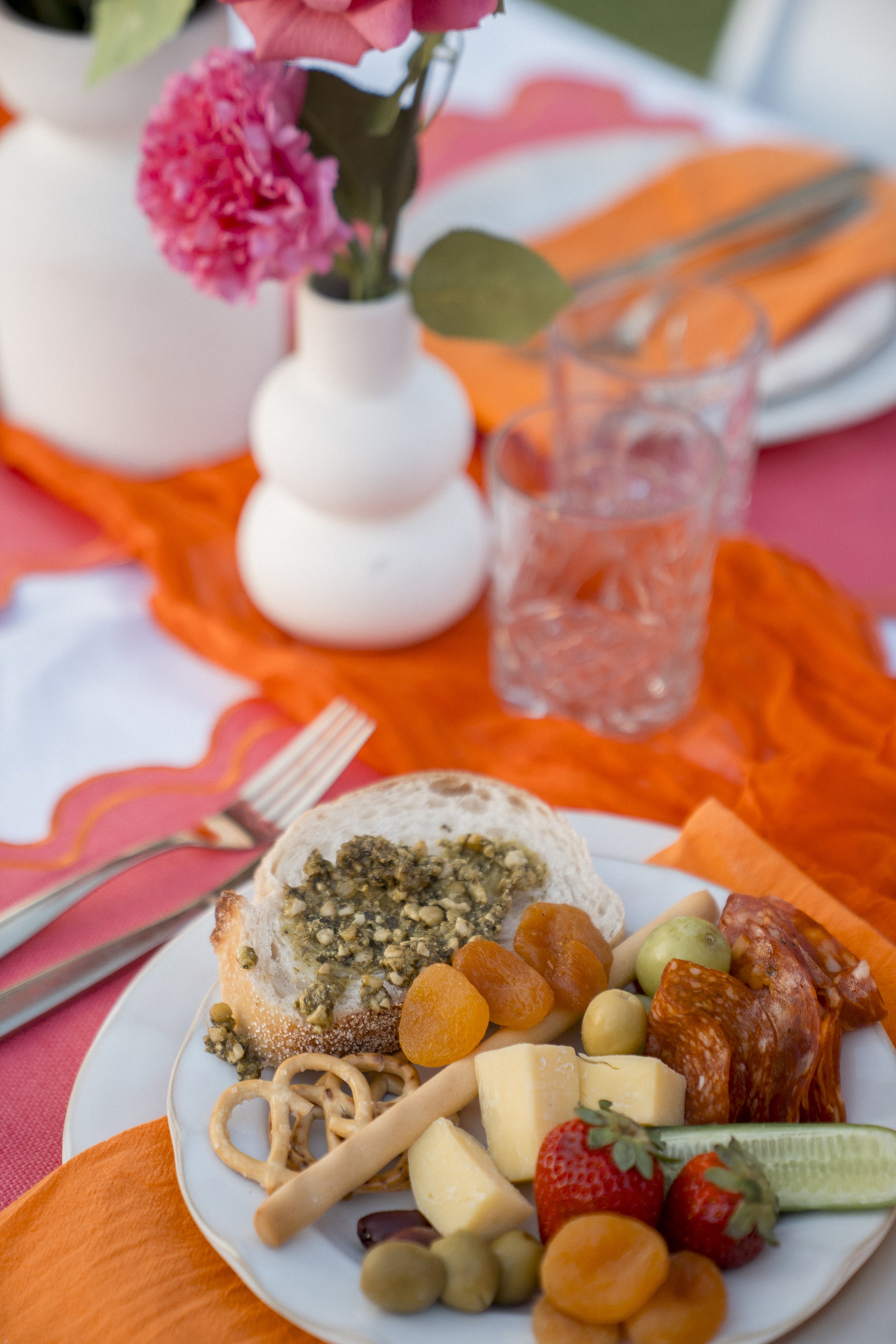 A table setting featuring a white plate with assorted cheeses, crackers, and fruits, a slice of bread with pistachio topping, and a glass of water. In the background, there are pink flowers in a white vase, orange napkins, and a fork on a pink and or