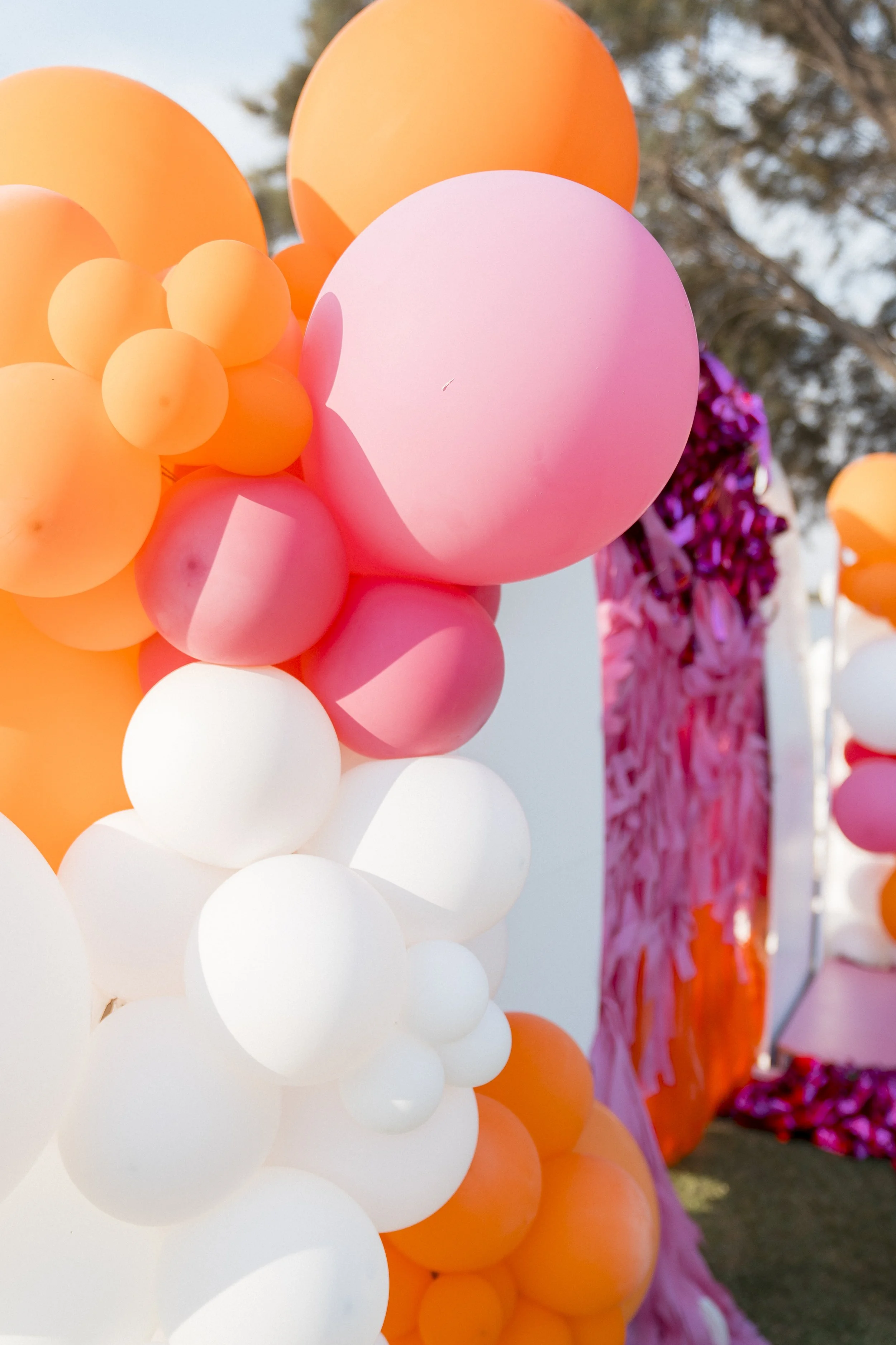 Colorful cluster of balloons, primarily in pink, orange, and white, with some purple decorations in the background outdoors.