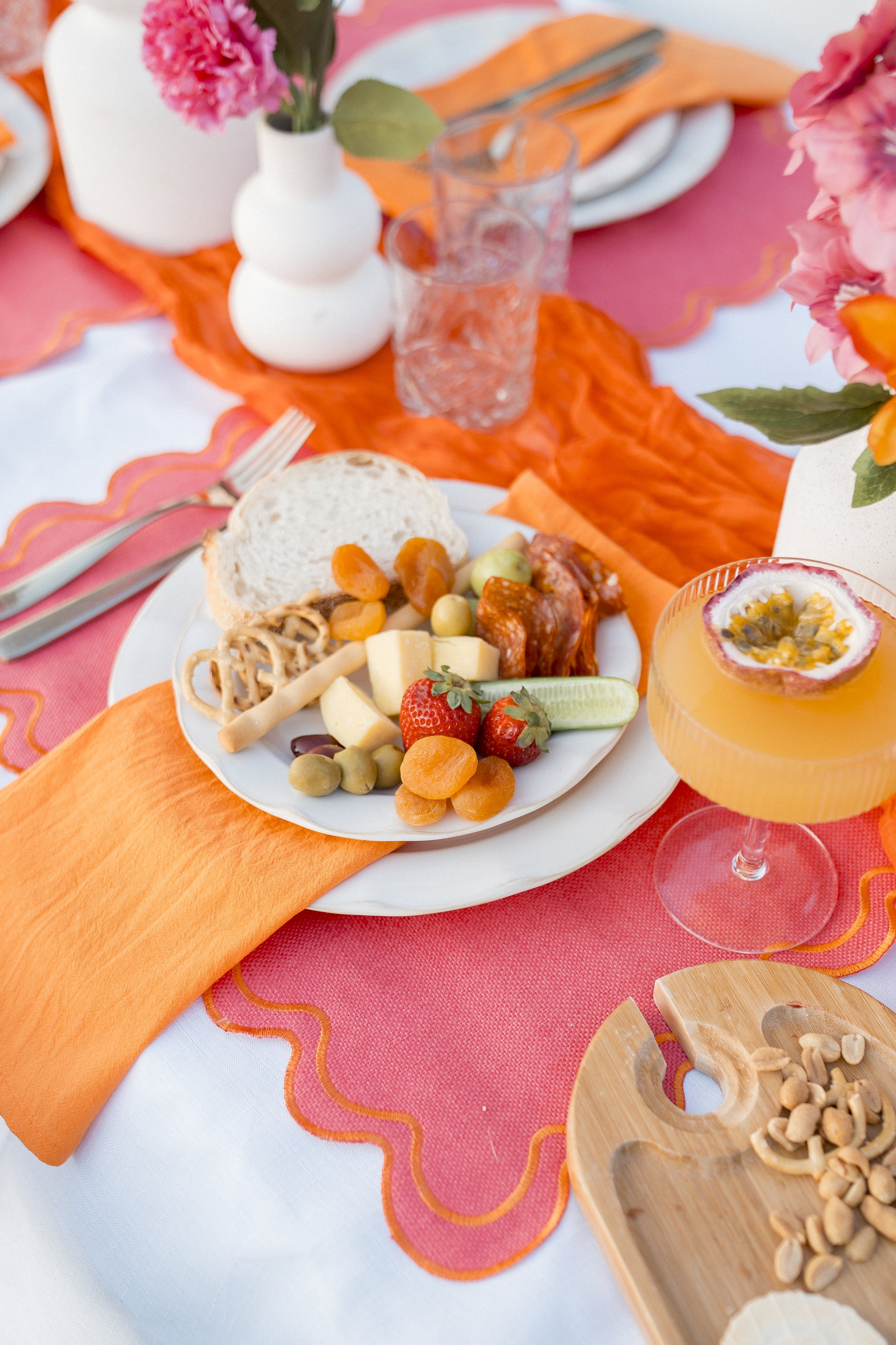 A table set for a meal with a pink and orange theme. There is a white plate with assorted cheeses, fruits, and nuts. A glass of orange beverage with passion fruit is in the foreground. The table has an orange table runner, pink napkins, and white pla