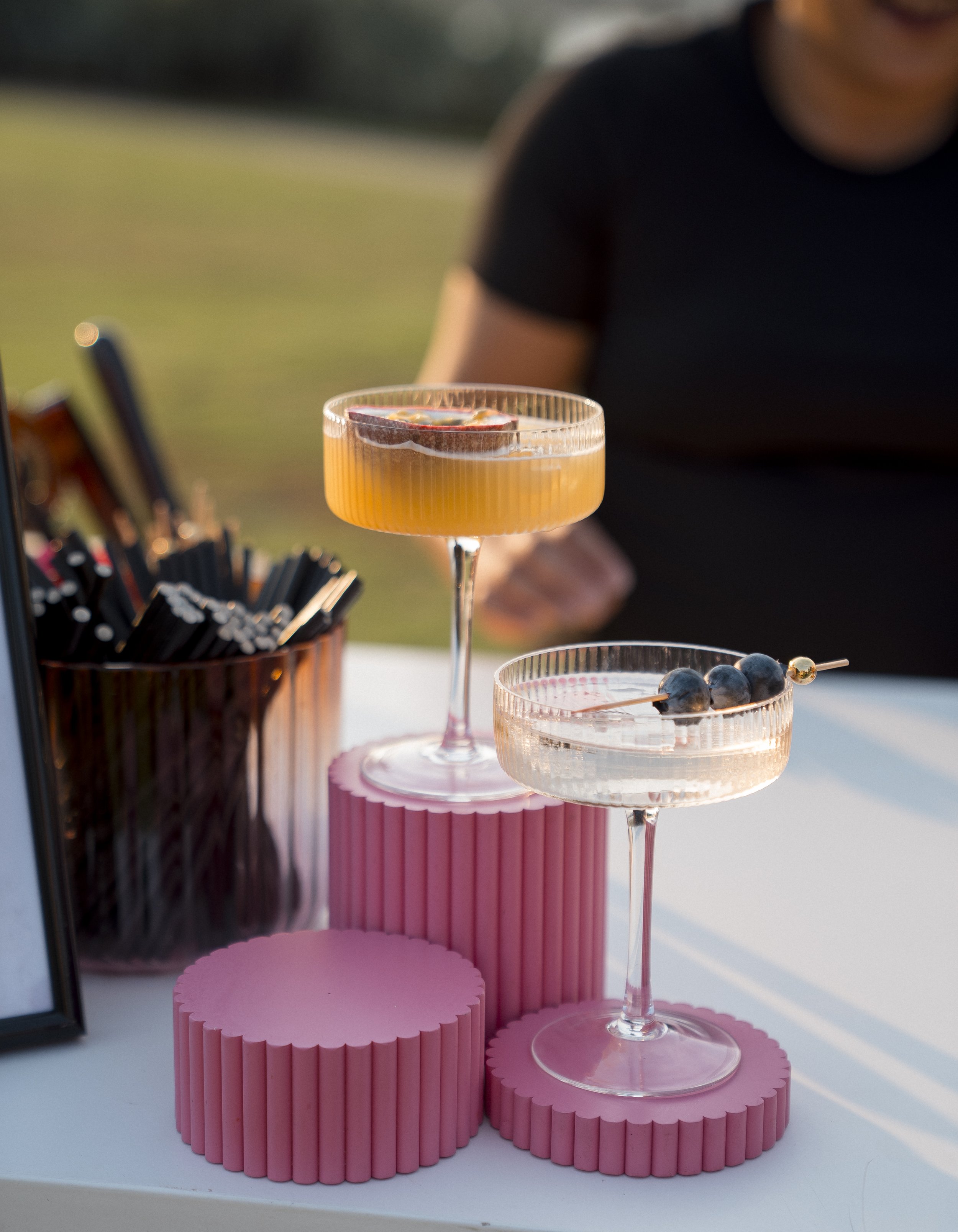 A pink fluted cake stand with two pink textured cake bases, holding two cocktails with garnishes, and a container with black straws nearby, on a white surface outdoors during daytime.