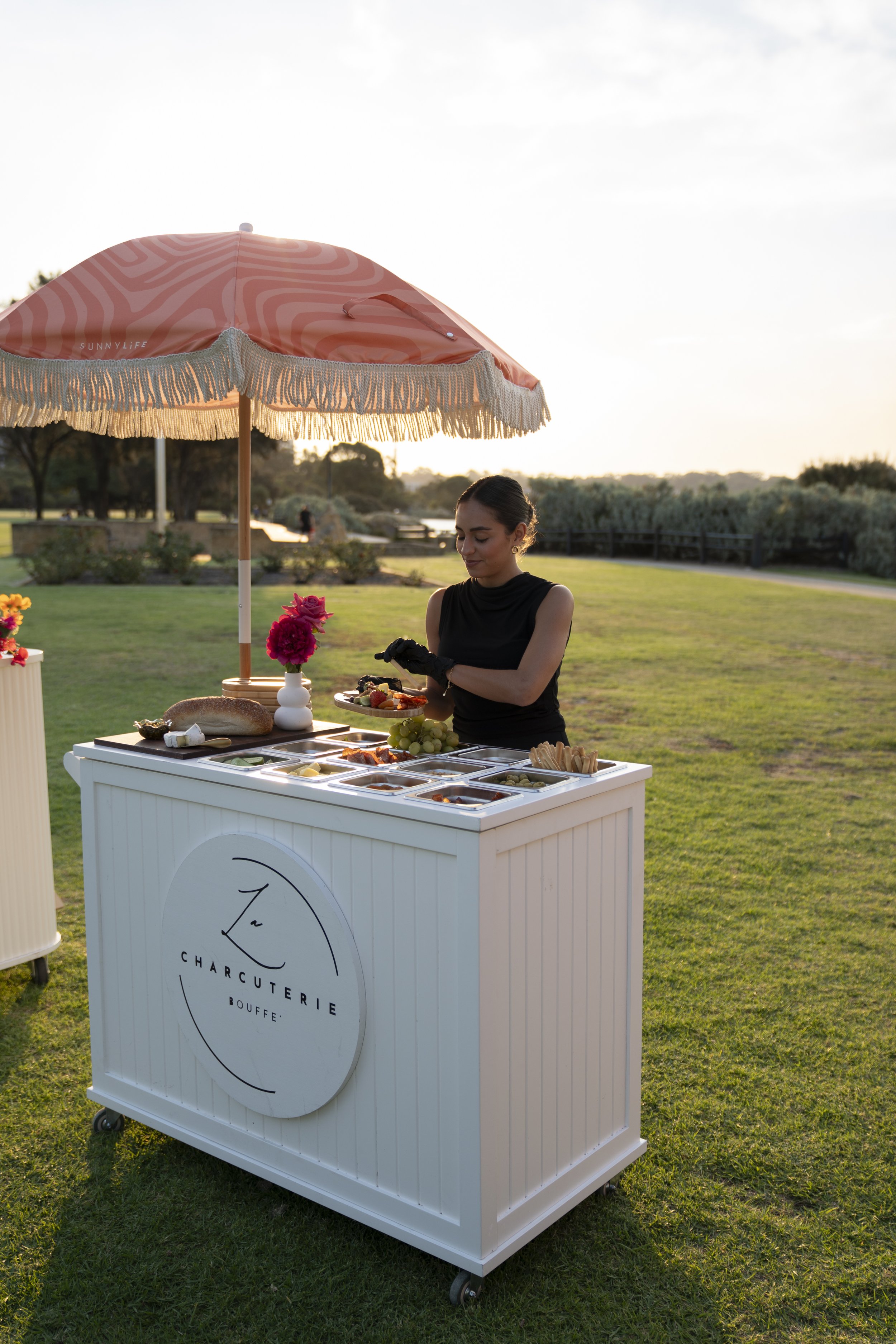 A woman standing outside under a pink umbrella with white fringe, serving herself at a white food station with an assortment of cheeses, bread, and grapes, on a grassy area during sunset.
