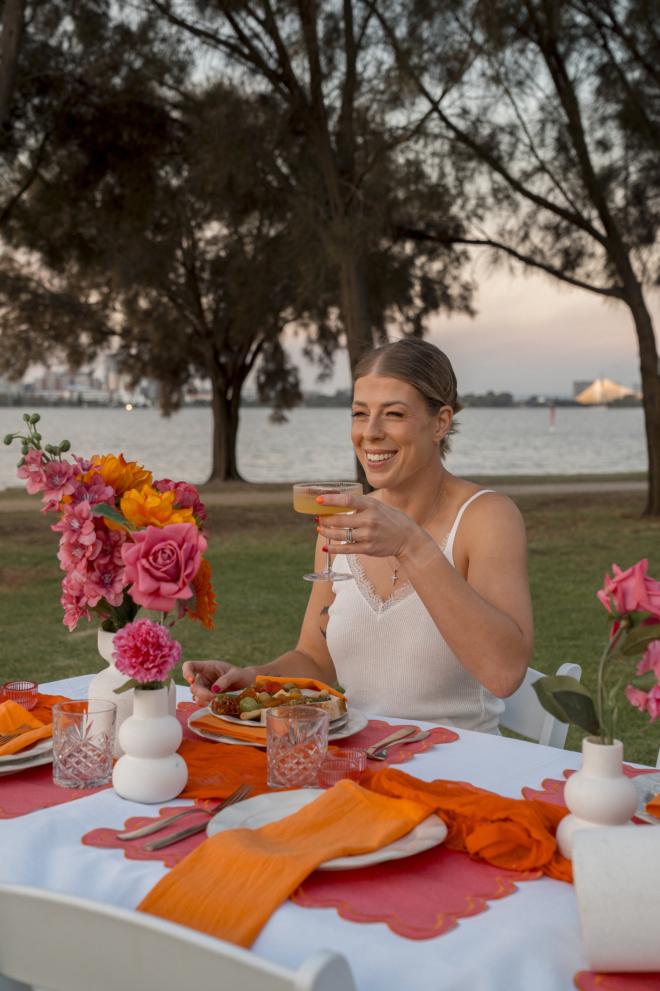 A woman with light skin, long hair, and a white tank top is smiling and holding a glass at an outdoor dining table with pink, orange, and white tableware, colorful flowers, and orange napkins. The setting is by a river with trees and a cityscape in t