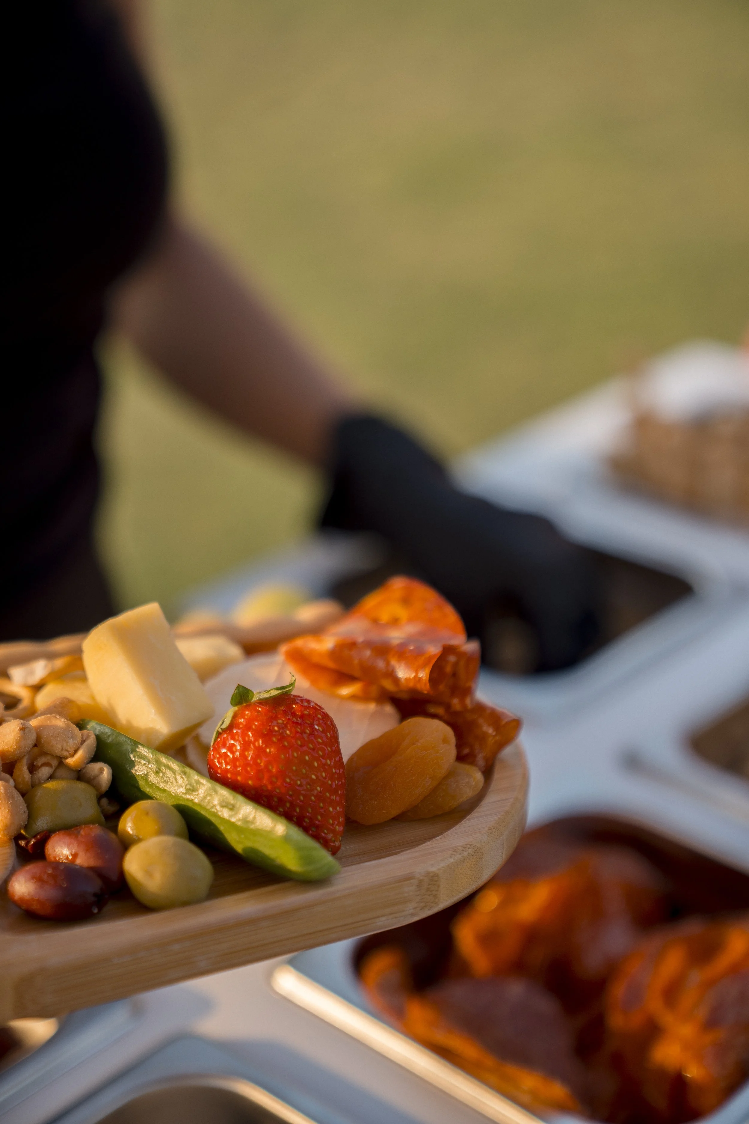 A wooden serving tray with assorted snacks including cheese, strawberries, nuts, chips, and pepperoni slices, with blurry dishes in the background.
