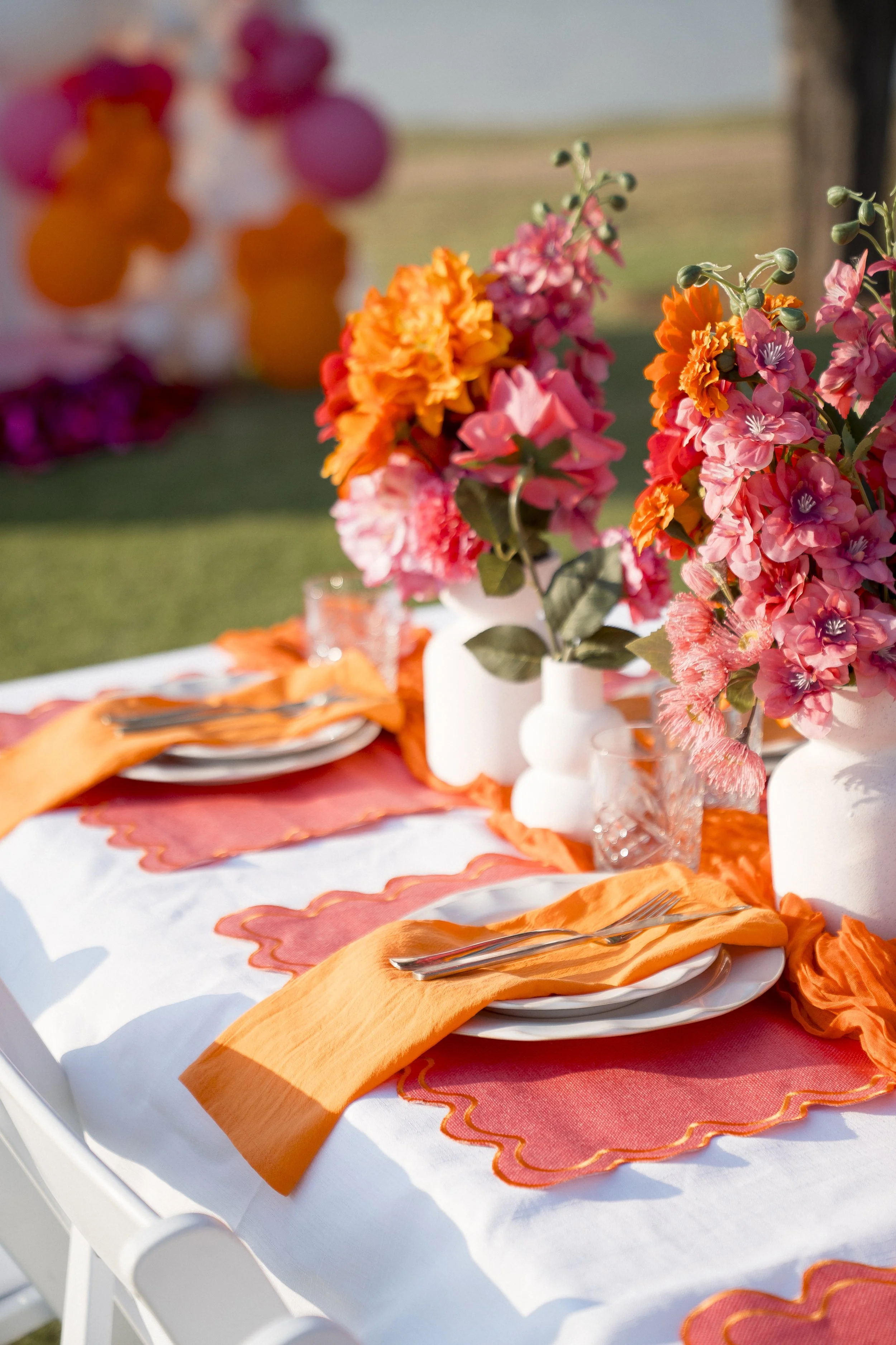 A table decorated with vibrant pink, orange, and purple flowers in white vases, set with white plates, orange cloth napkins, and silverware, on a white tablecloth with orange and pink scalloped placemats, outdoors on a sunny day.