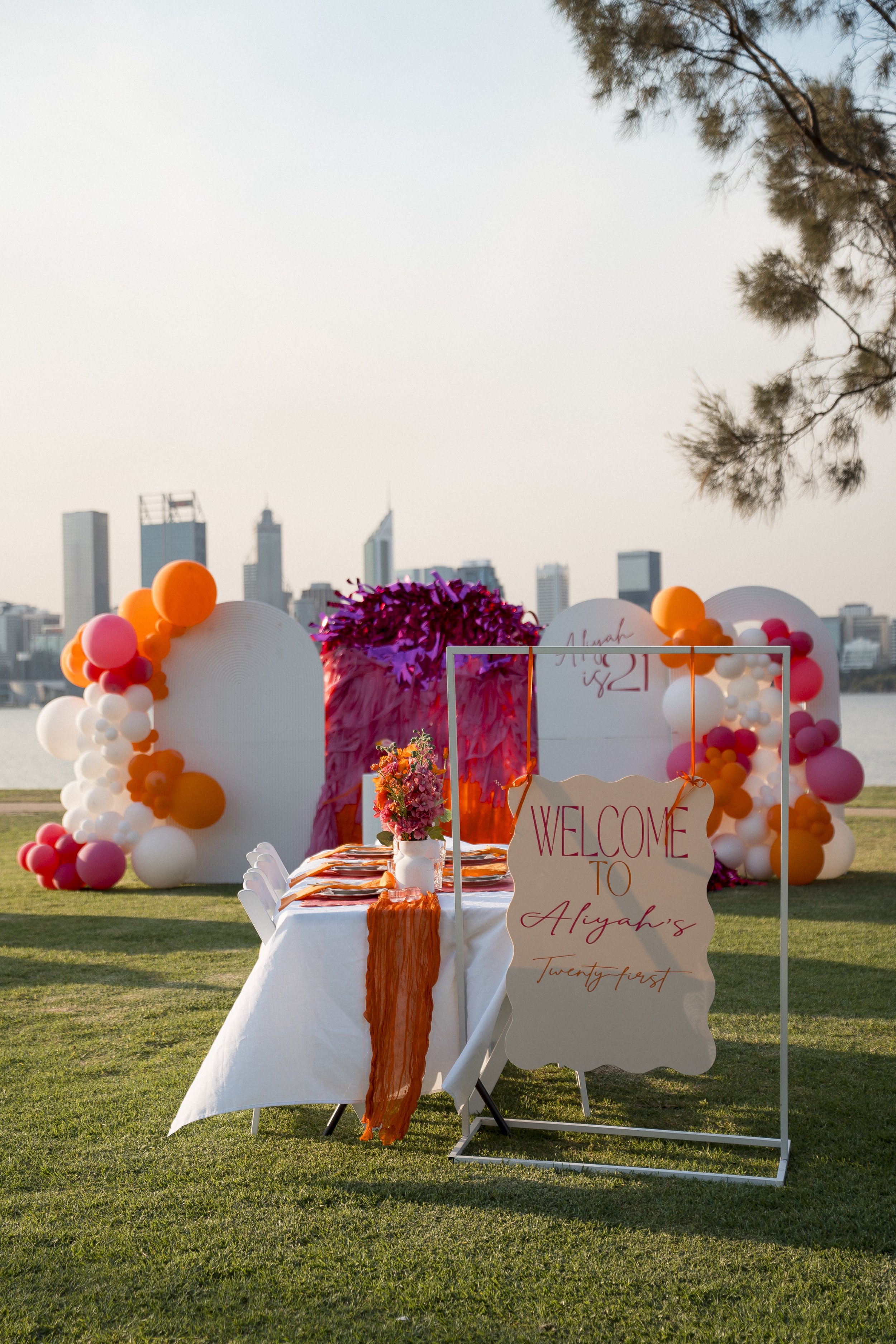 Decorations for a celebration or event on a grassy outdoor area, with a city skyline in the background, featuring balloons and a welcome sign.