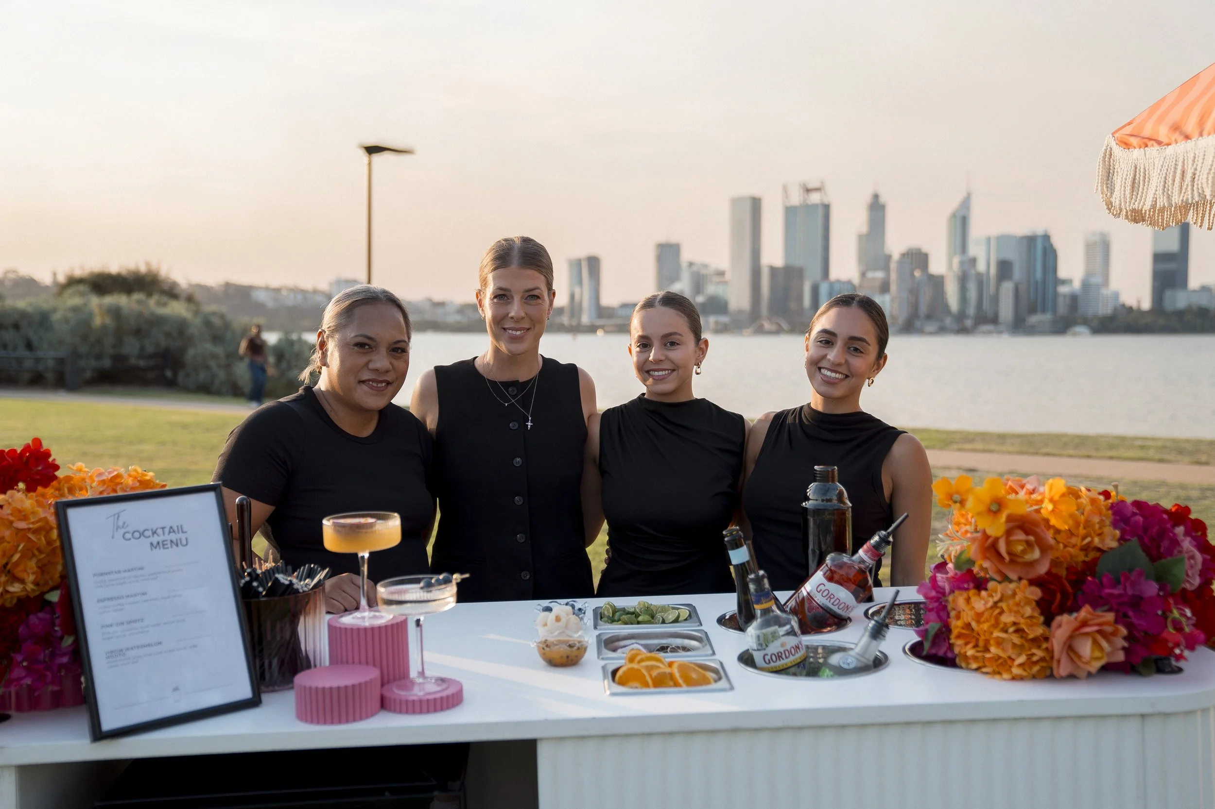 Four women standing behind a table with flowers, drinks, and a cocktail menu at an outdoor event by a body of water with a city skyline in the background.