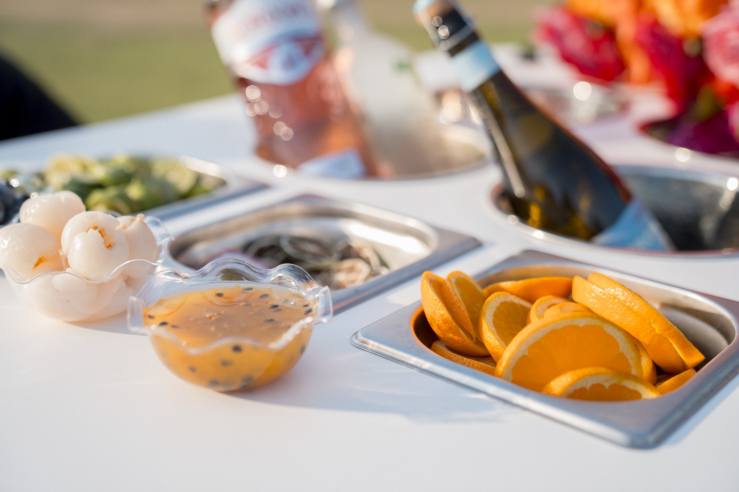 Fruits and sauces on a buffet table, including sliced oranges, a bowl of peeled lychee, and a small bowl of orange sauce with black seeds.