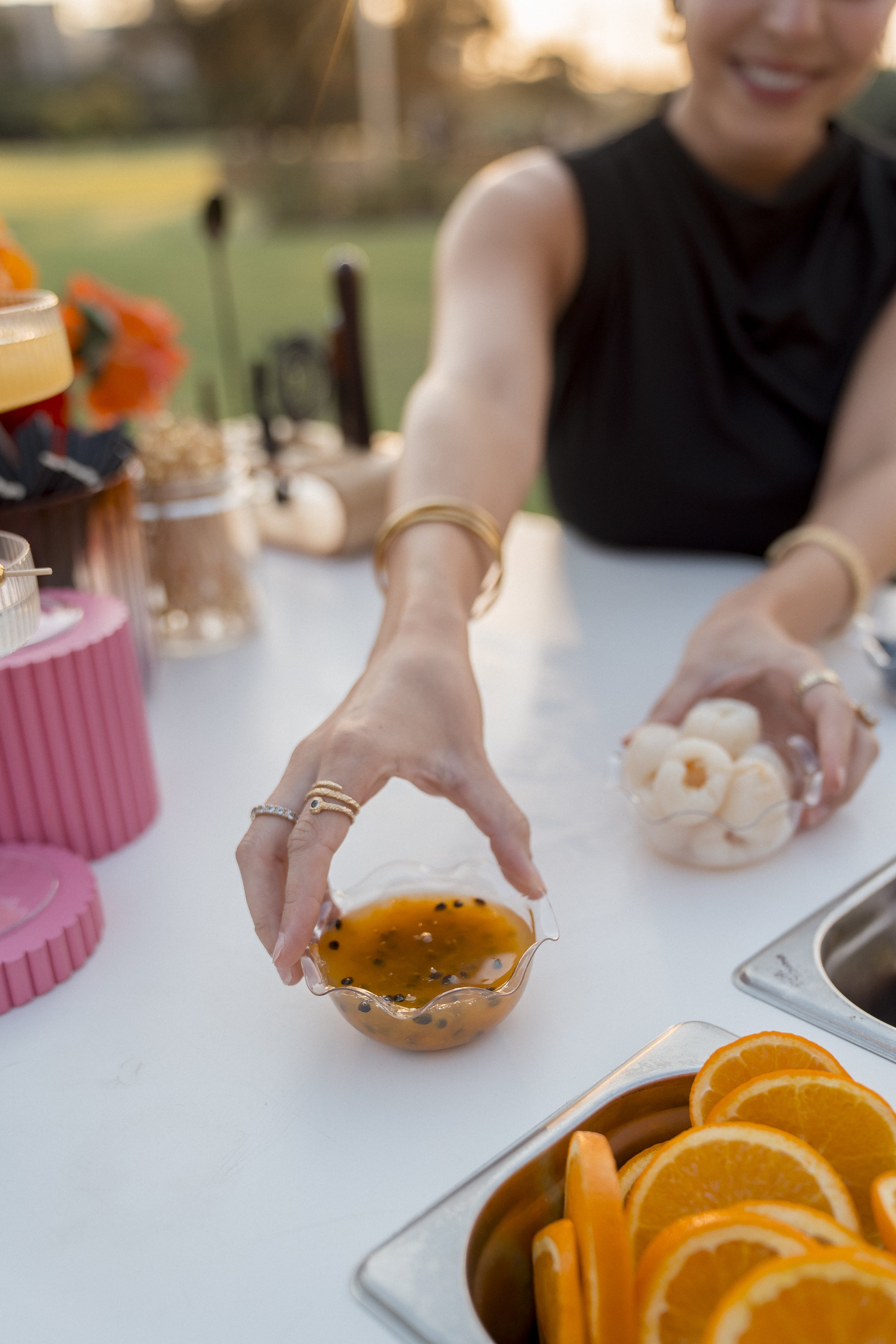People preparing fruit at an outdoor gathering, with a focus on hands reaching for a bowl of passion fruit and a tray of sliced oranges on a white table, with blurred background including flowers and rolling landscape.
