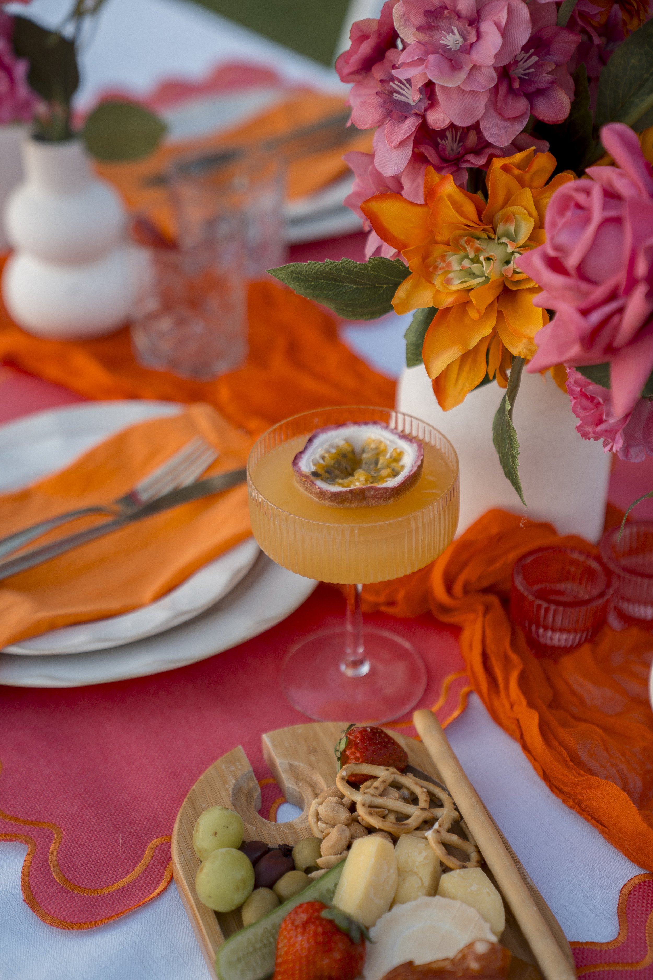 Colorful table setting with a bouquet of pink and orange flowers, a glass with orange-colored drink garnished with passion fruit, a cheese and fruit platter with strawberries, grapes, cheese, and crackers, and an orange tablecloth with decorative edg