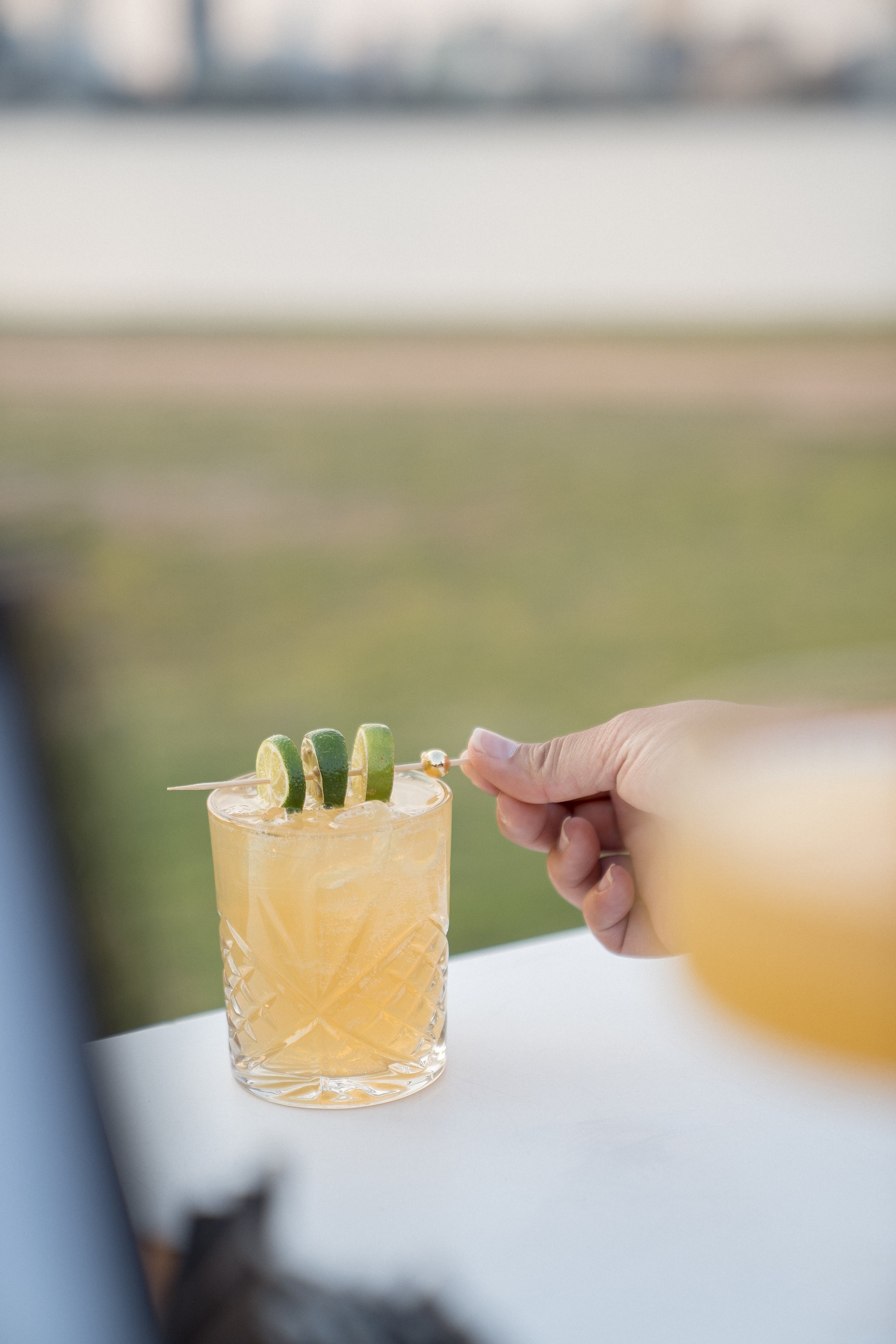 Close-up of a person holding a cocktail glass with a yellowish drink, garnished with a slice of lime and a small skewered piece of fruit or vegetable, with a blurred background of outdoor scenery.