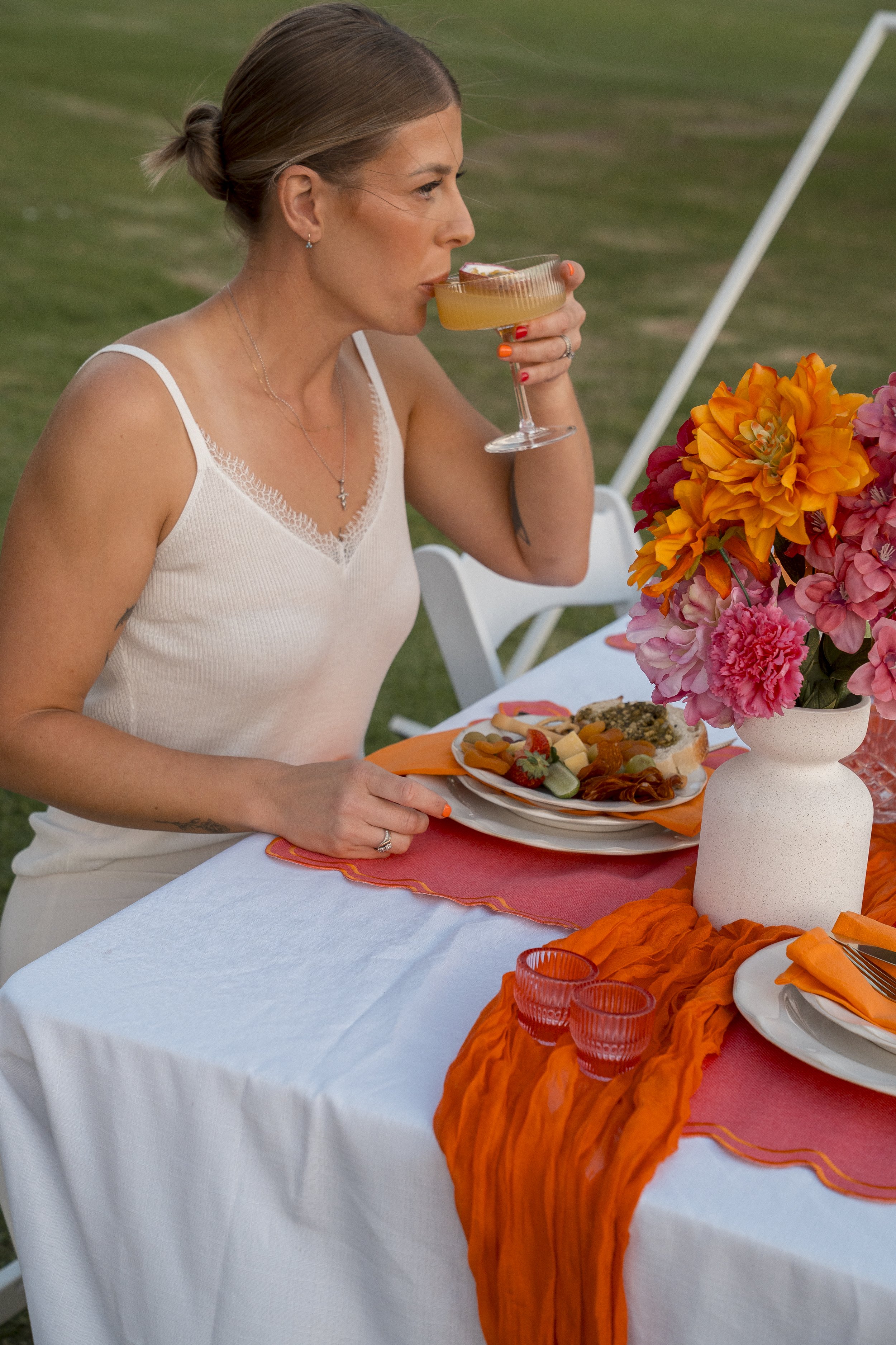 A woman with light skin, brown hair tied back in a bun, wearing a white tank top and jewelry, drinking from a cocktail glass at a decorated outdoor dining table with orange and pink floral arrangements, orange napkins, plates with food, and orange ta