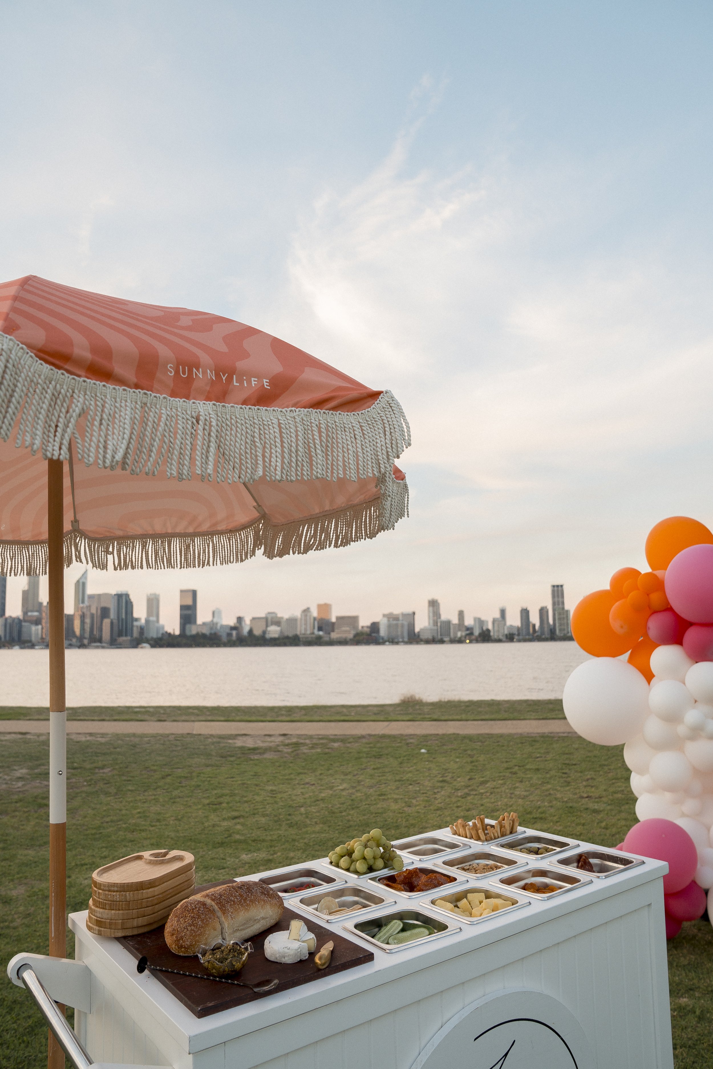 A street food cart with a white cooler containing cheese, bread, grapes, and various toppings, set outdoors near a lake with a city skyline in the background.