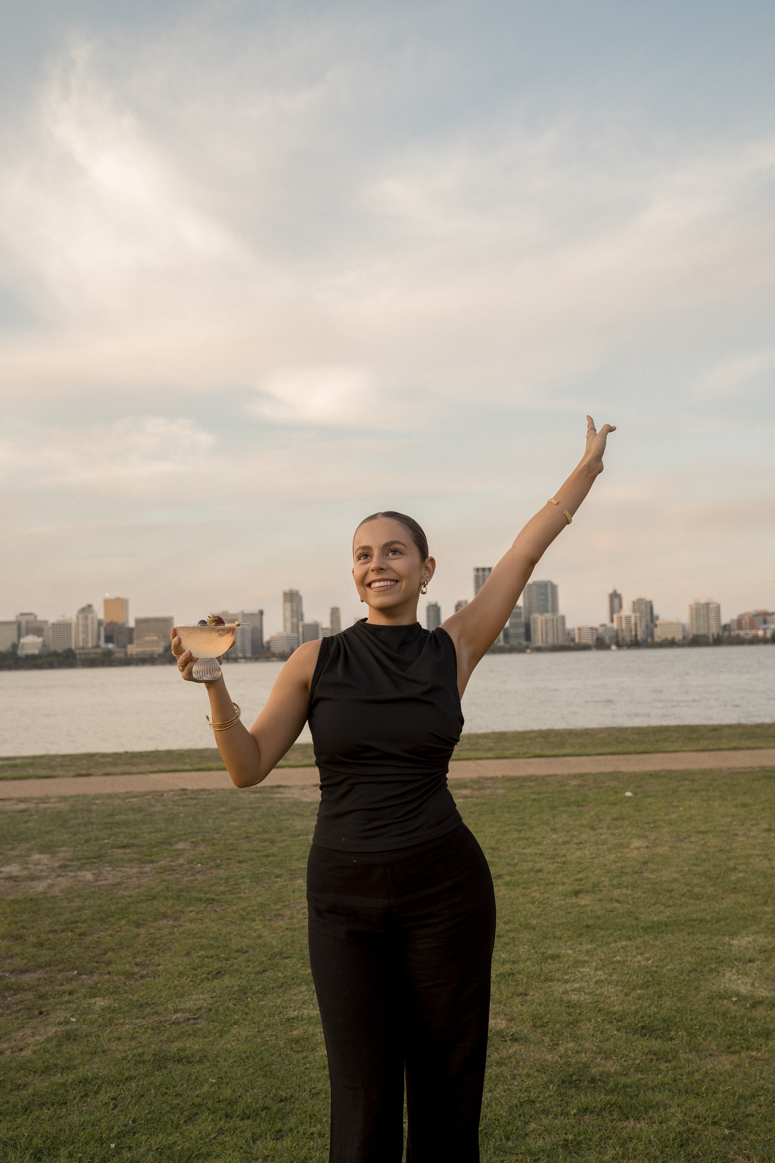 A woman in a black dress standing outdoors by a river with a city skyline in the background. She is smiling, holding a cocktail in her right hand, and raising her left arm in the air.