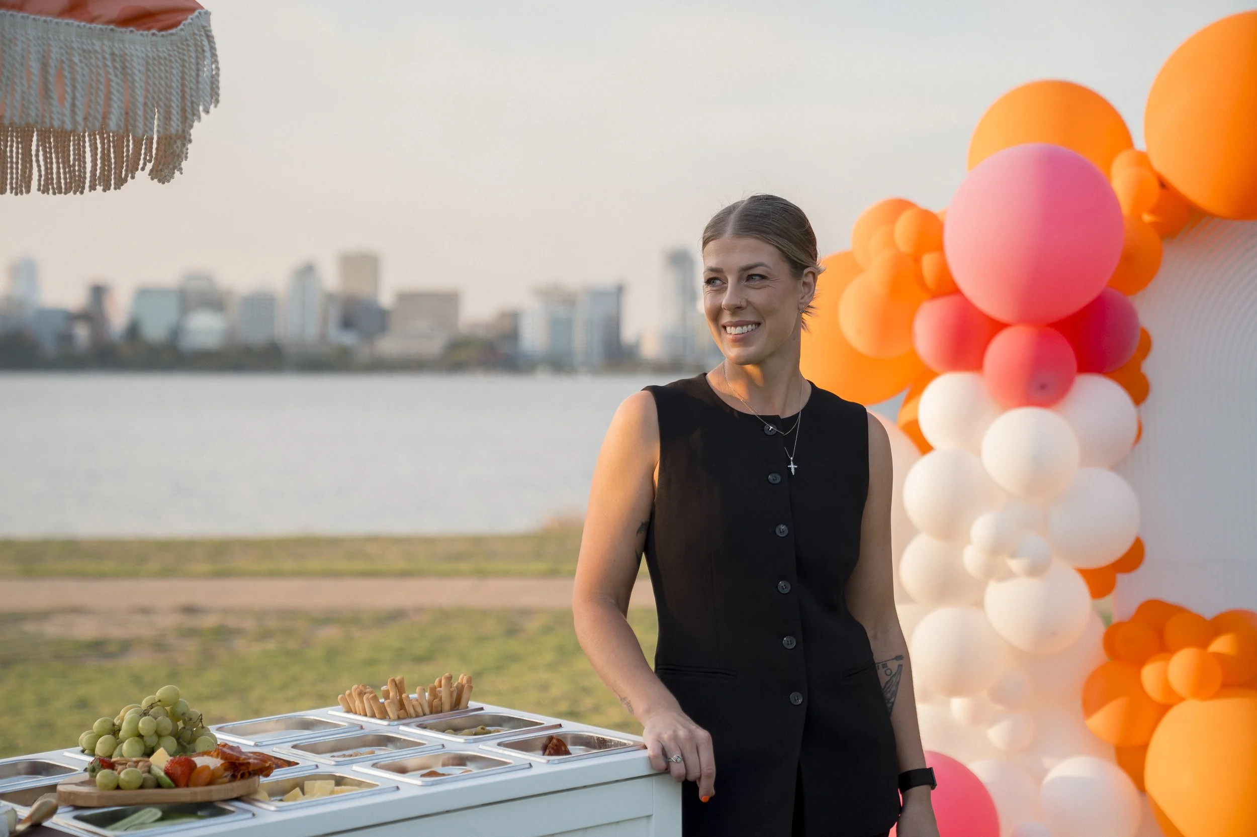A smiling woman with a black dress and layered necklaces stands outdoors near a table with various food items and cheese and grapes. Behind her, there is a colorful balloon decoration with pink, orange, and white balloons, and a city skyline across a