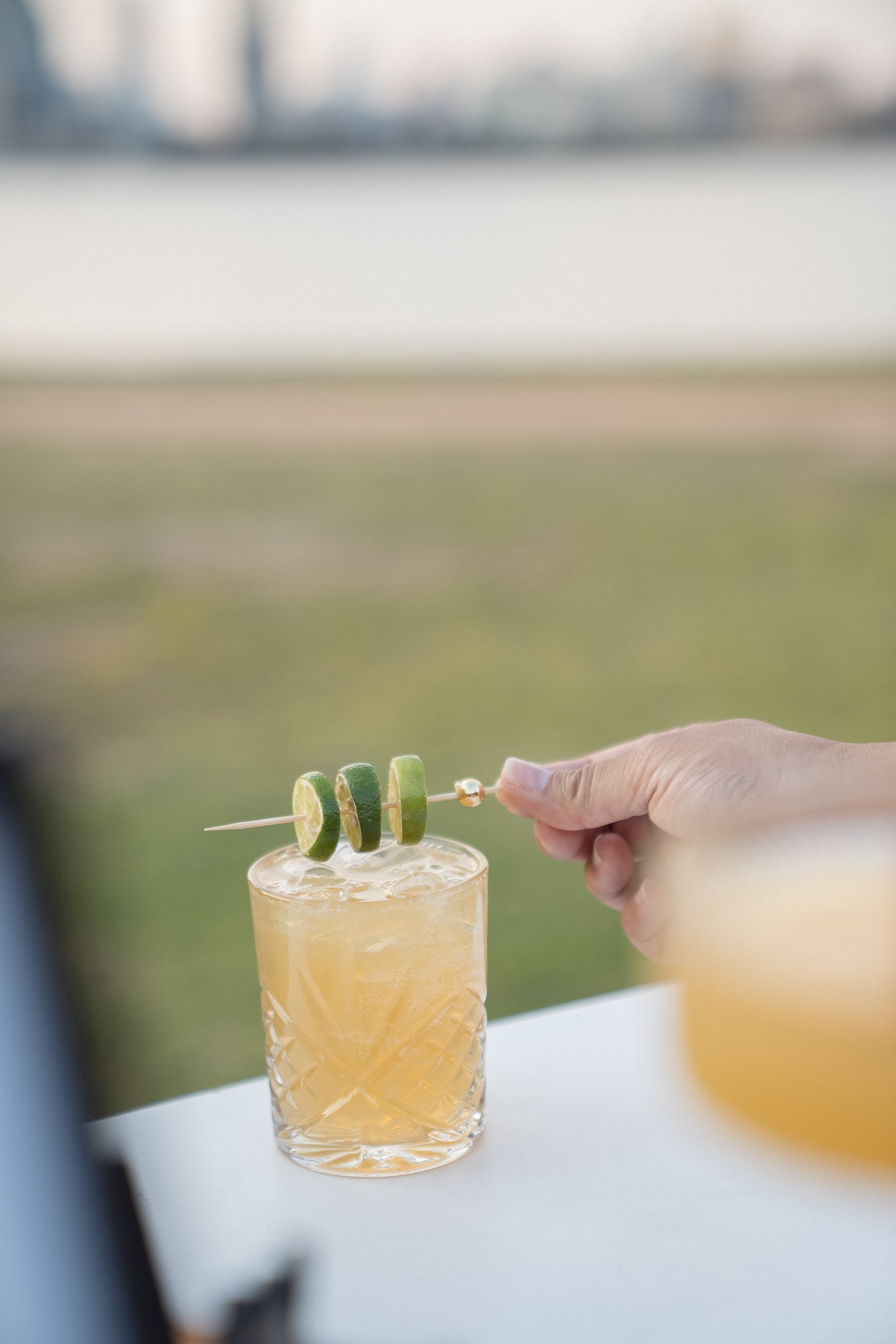 A hand placing a skewer with slices of cucumber into a glass filled with a light orange beverage, set outdoors with a blurred background.