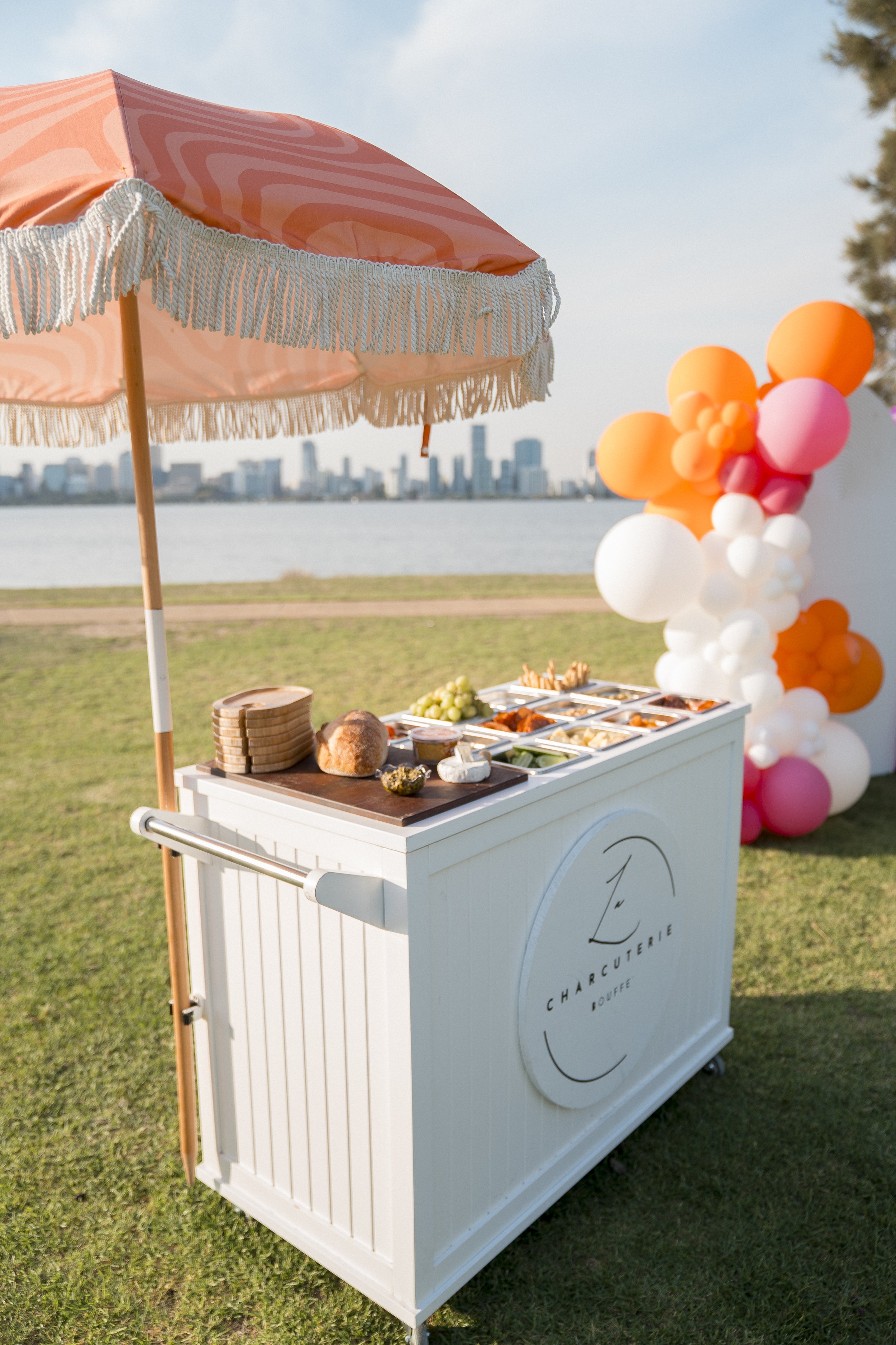 A white catering cart with a curved logo that reads 'Charcuterie' and 'Louffre' underneath, set on grass near a body of water with a city skyline in the background. The cart has a large pink and orange balloon bouquet attached to it, and a table on t