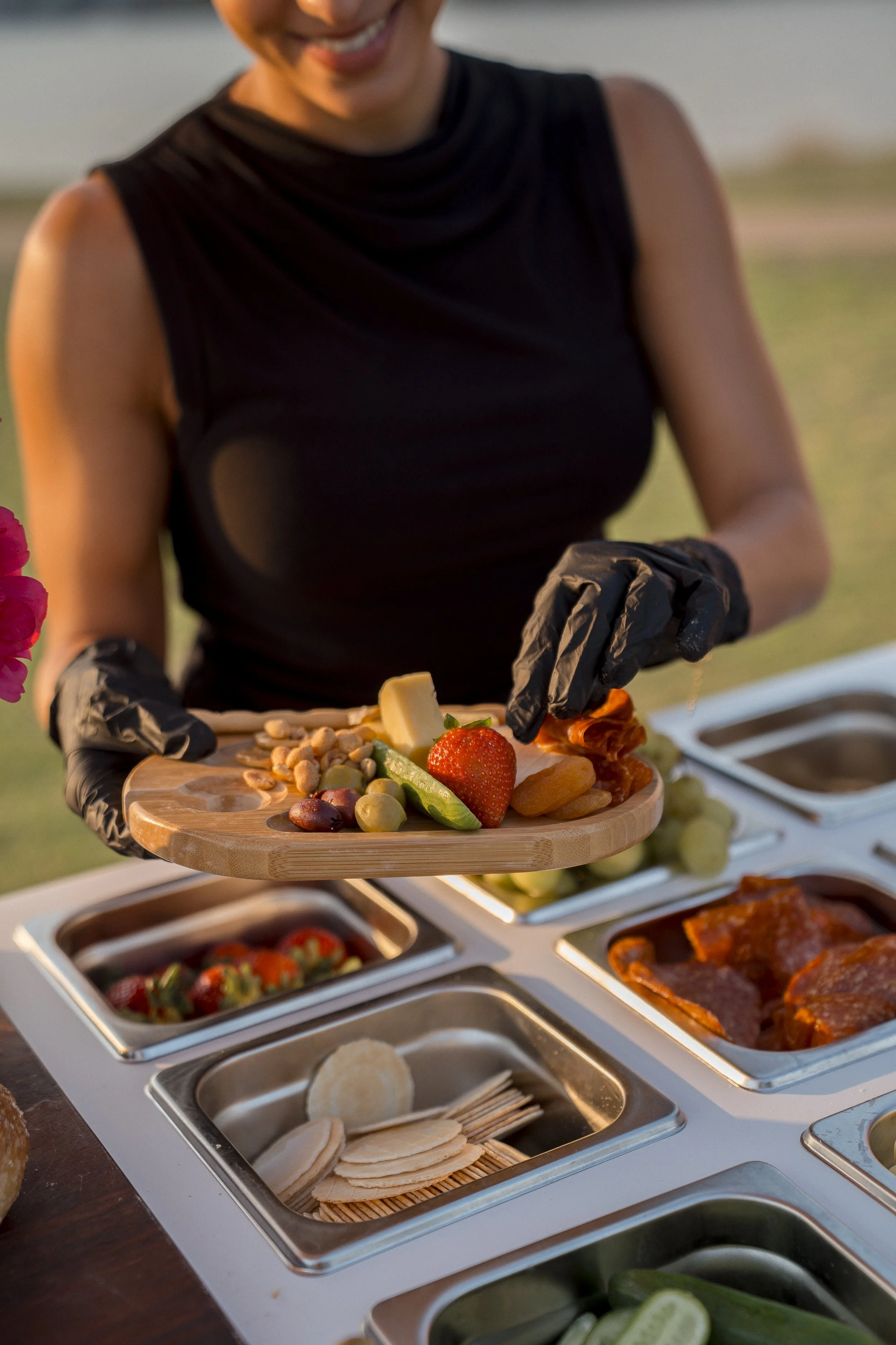 A person wearing a black sleeveless top and black gloves serving a selection of food on a wooden platter, with a buffet of various dishes including strawberries, grapes, cheese, and crackers in front of them.