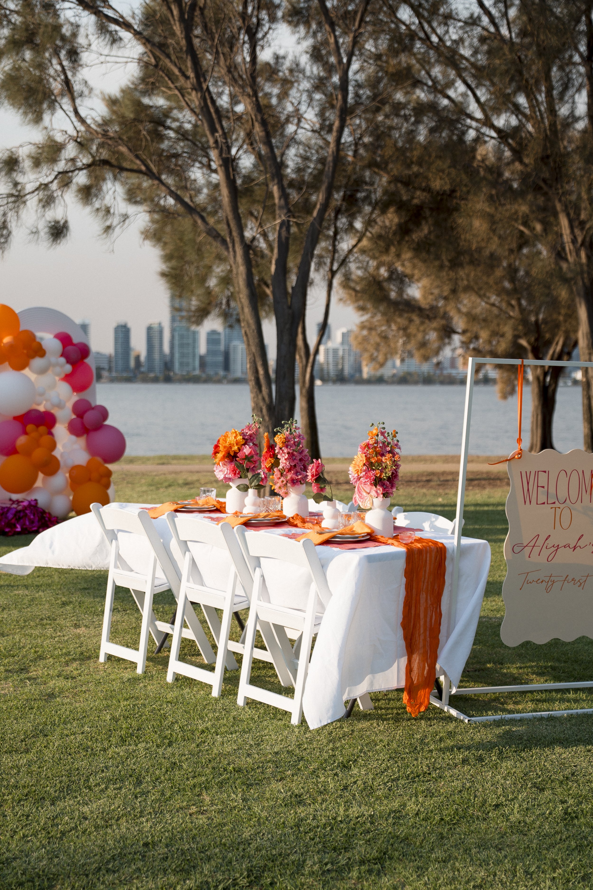 A decorated outdoor table setup with pink, orange, and white flowers, orange table runner, and pink napkins, with a balloons arch in pink, orange, and white on the left side, overlooking a river with city skyline in the background, trees framing the 