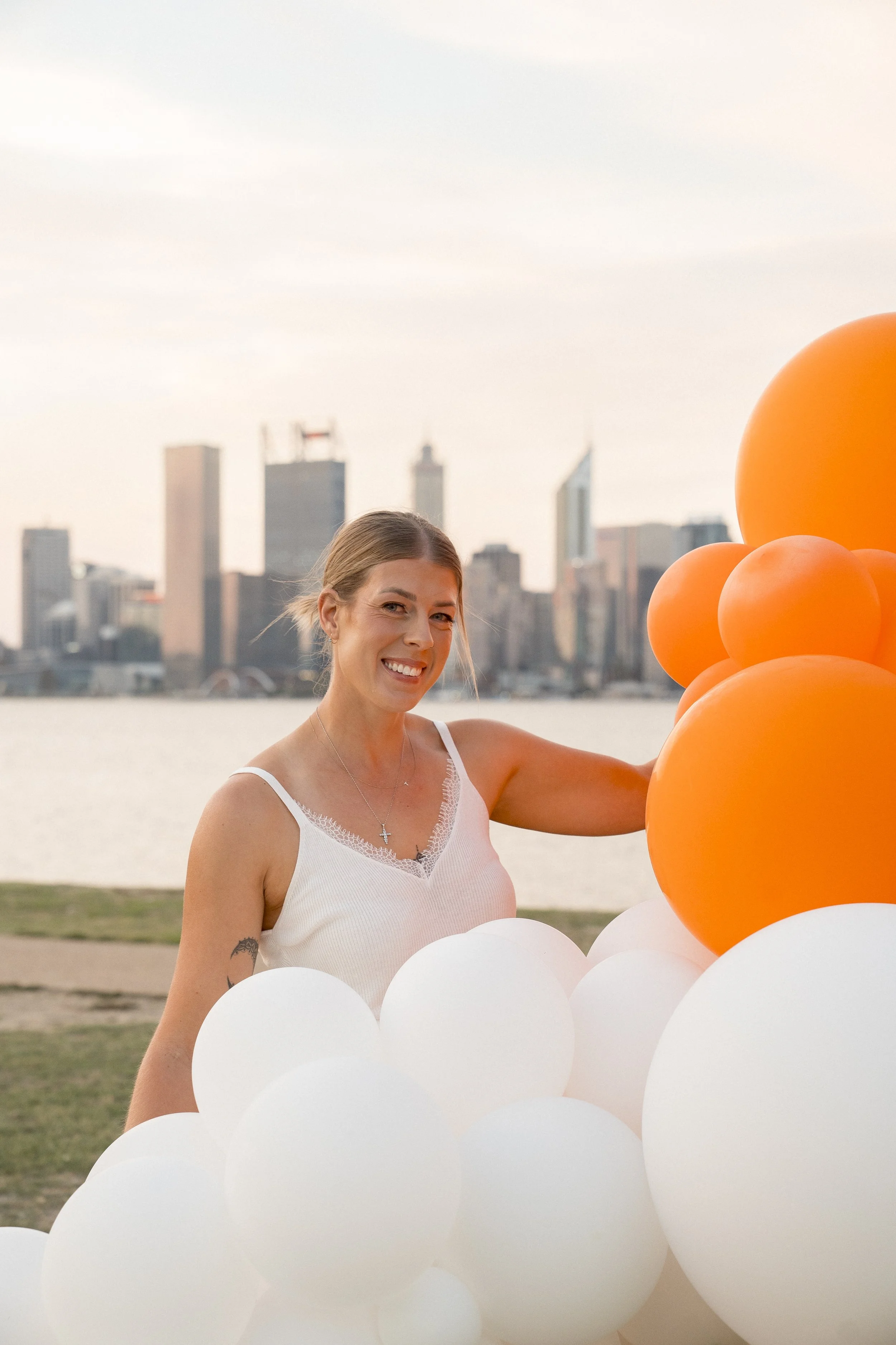 A woman smiling outdoors holding a bunch of white and orange balloons with a city skyline in the background during sunset.