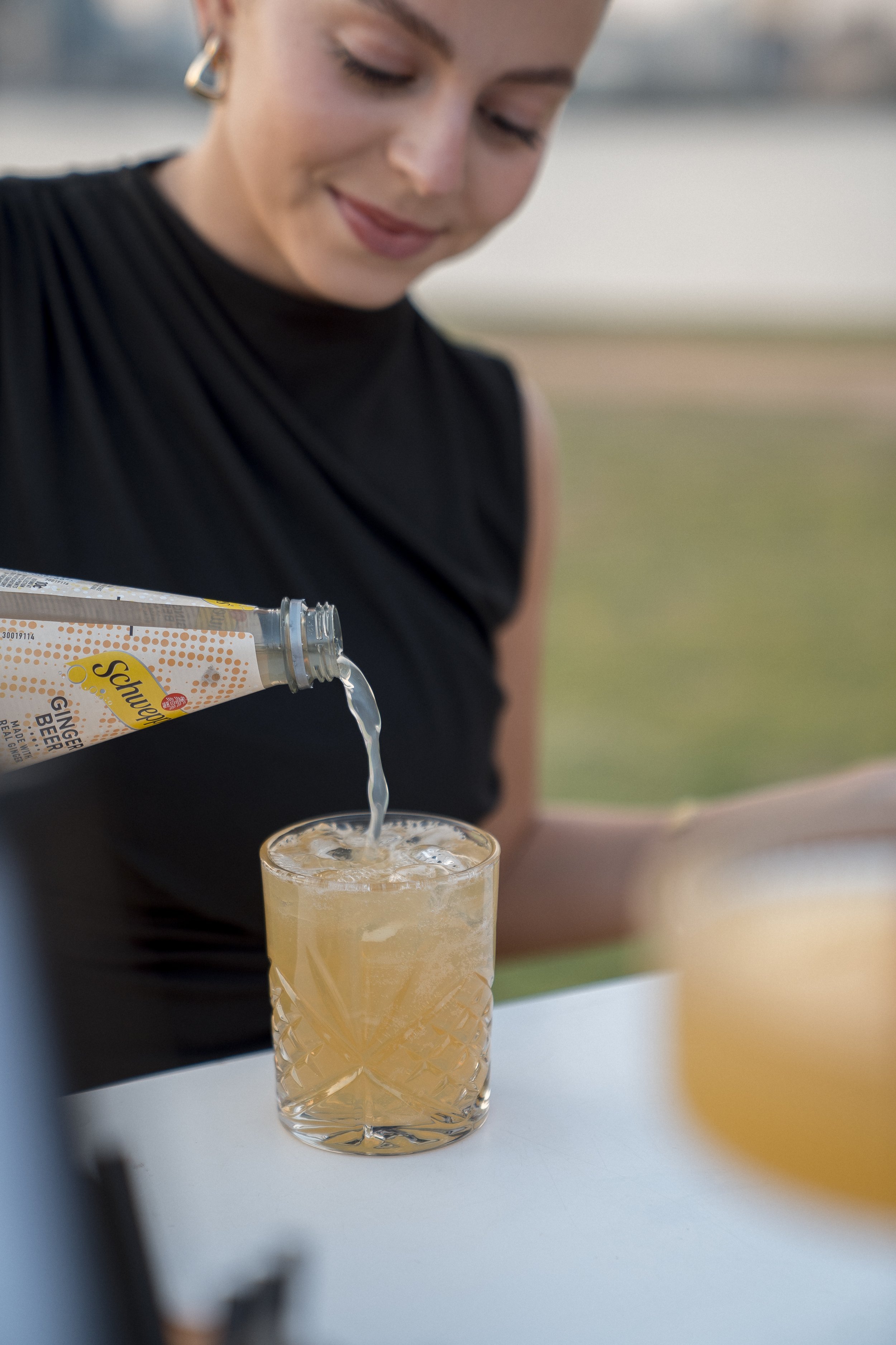 A woman in a black sleeveless top sprinkles gin from a Schweppes ginger beer bottle into a textured glass filled with ice, outdoors during daytime.