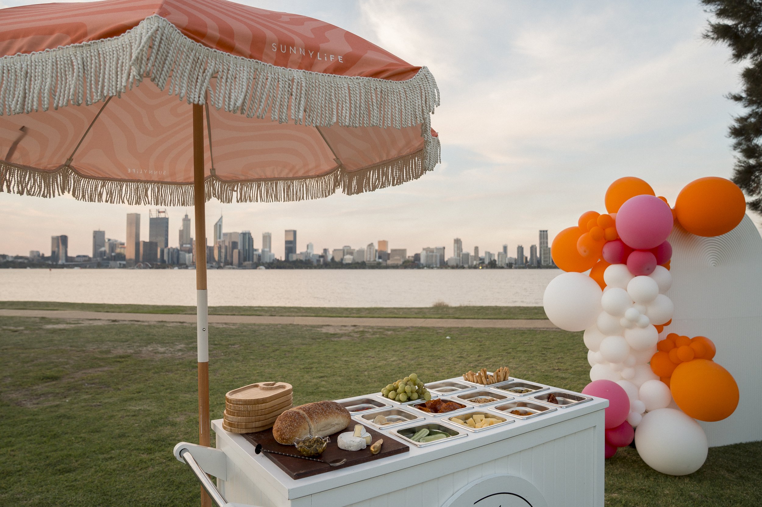 A food stand with a hat of bread on a checkout cart with salads and cheeses, and surrounded by a colorful balloon decoration during sunset with a city skyline visible in the background.
