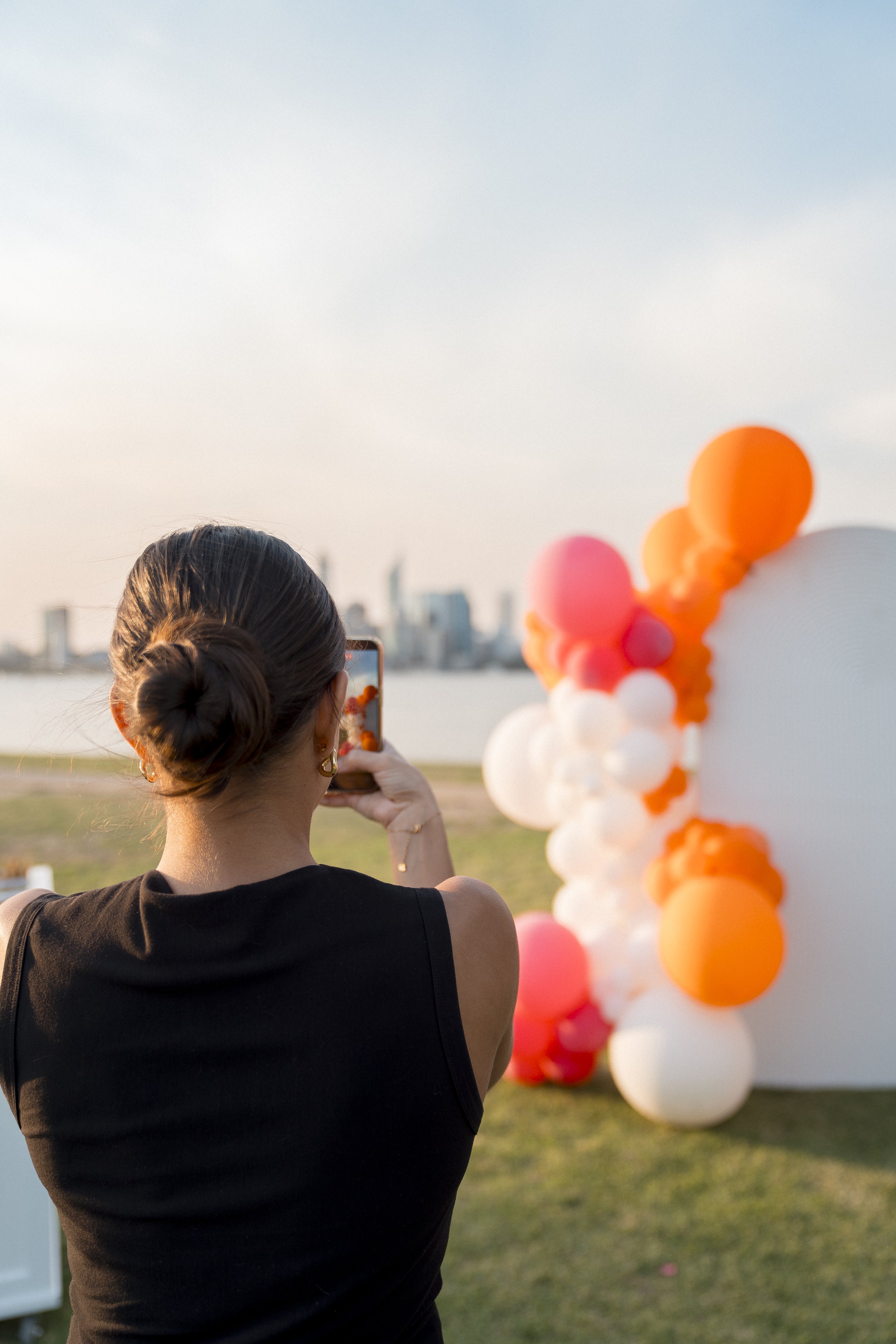 A woman with dark hair in a bun is taking a photo or video with her smartphone of a balloon arrangement with pink, orange, and white balloons at an outdoor event during sunset, with a city skyline in the background.