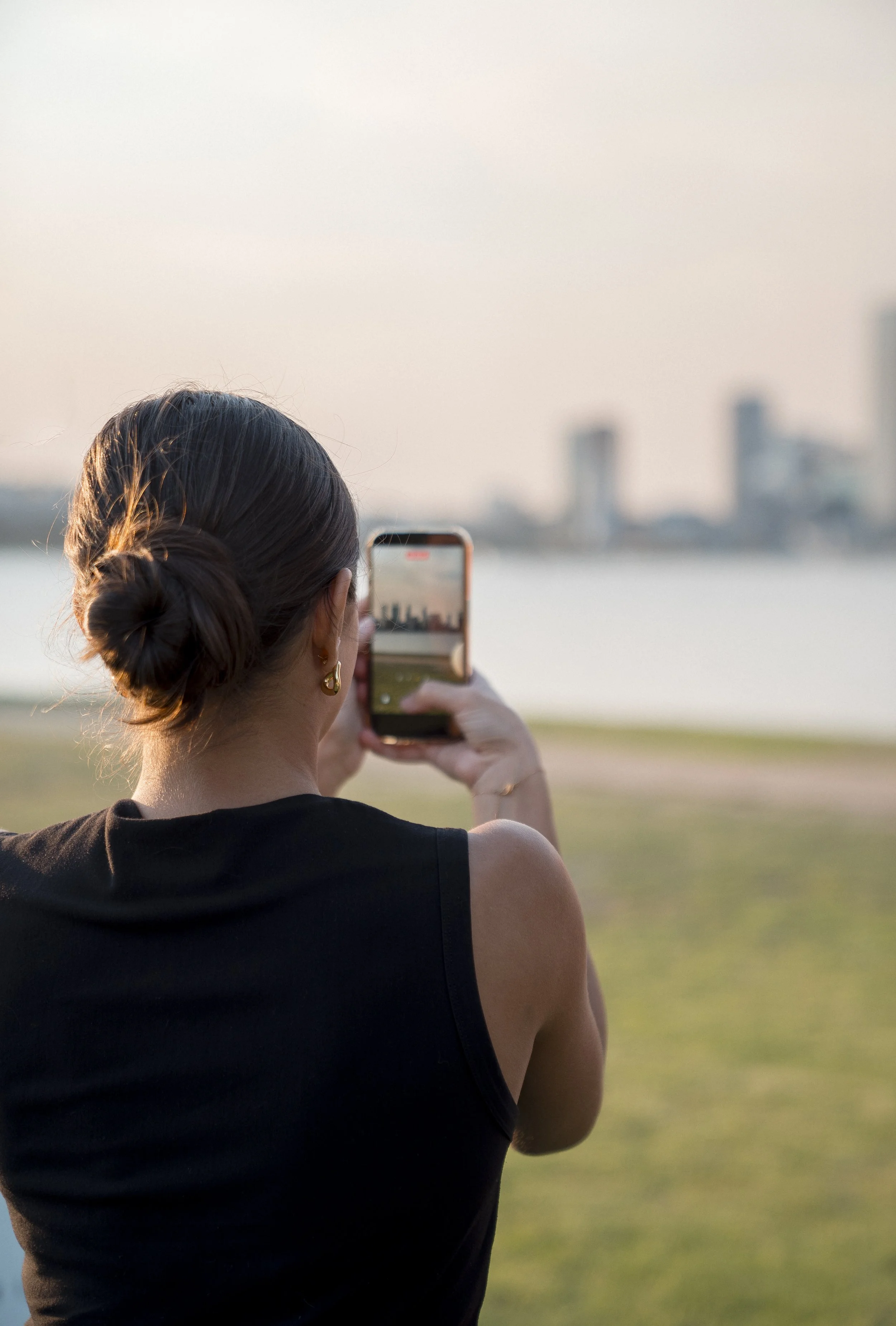 Woman with brown hair tied back in a bun, wearing earrings, taking a photo of a city skyline with her smartphone in a park during sunset.