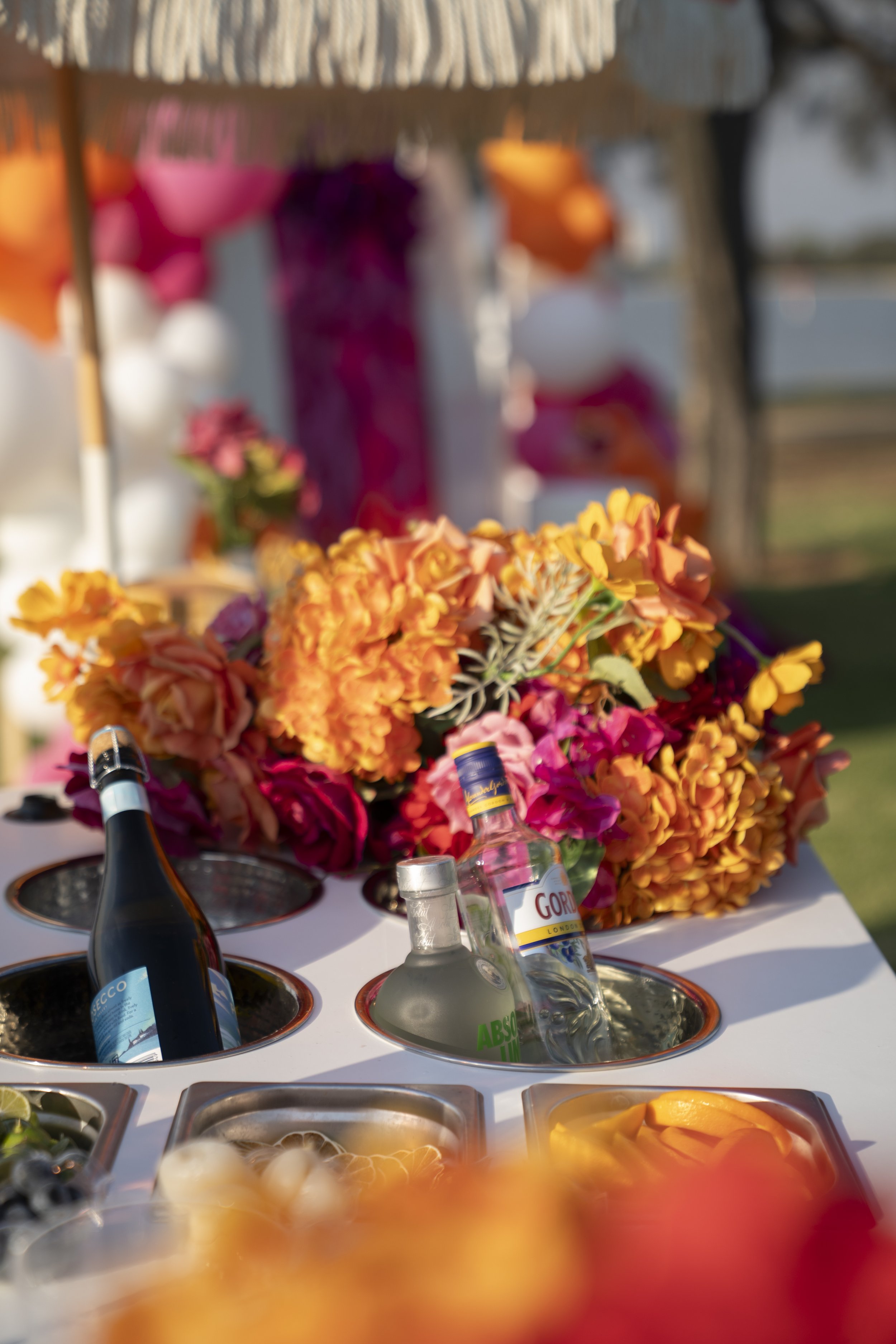 A table with a colorful flower arrangement, bottles of alcohol, and food items, likely at a celebration or outdoor event.