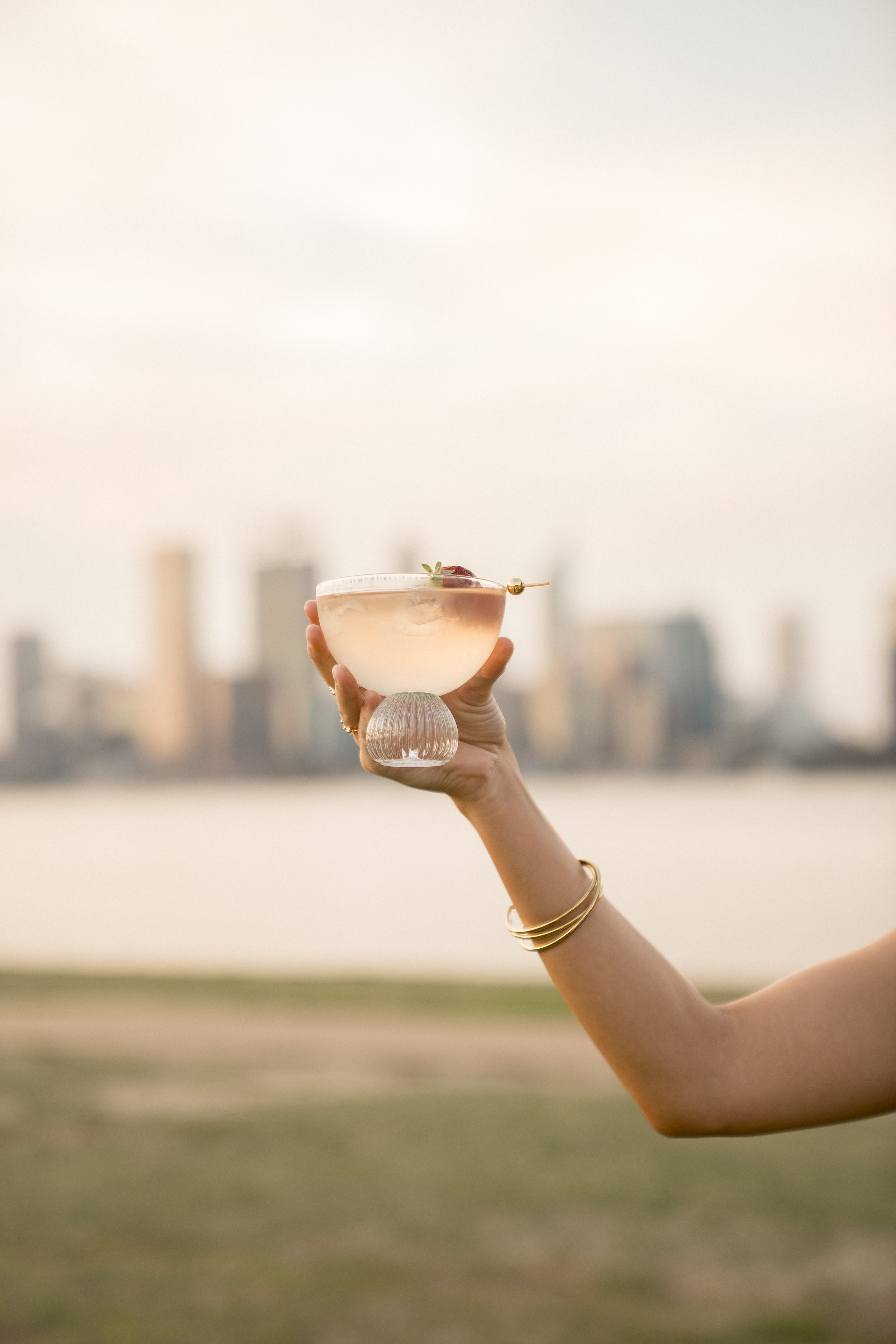 A hand holding a glass of pink cocktail with a small skewer garnished with a strawberry, against a blurred city skyline background during sunset or dusk.
