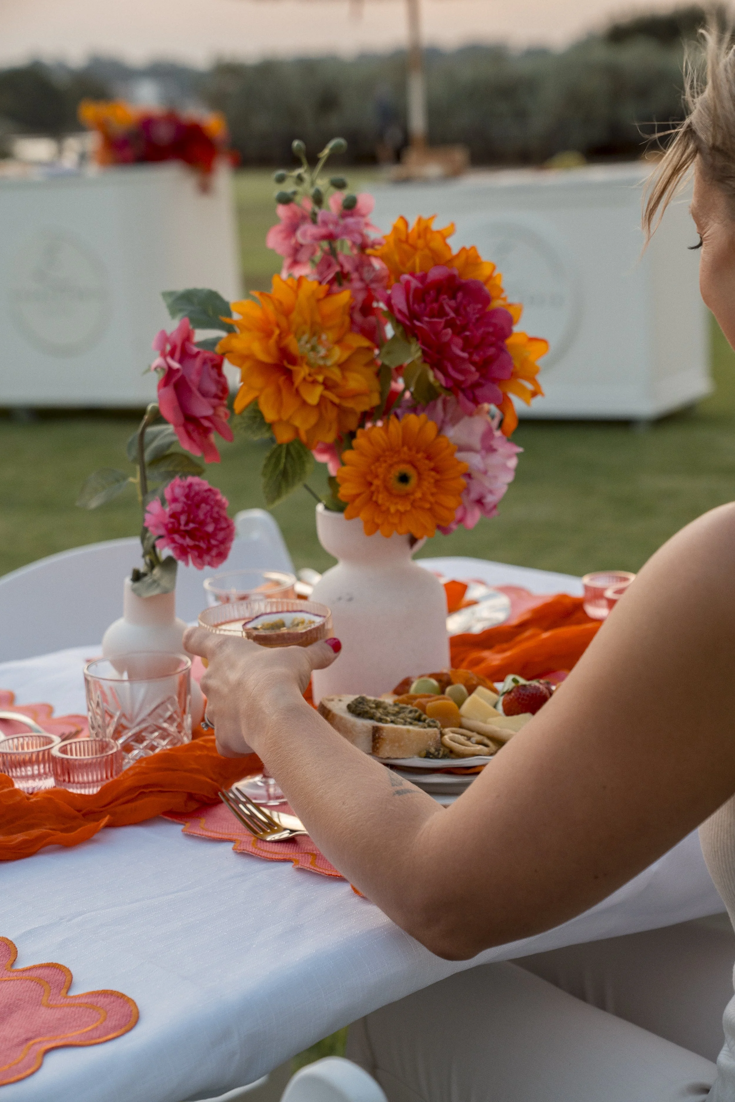 A person holding a glass of tea or wine at an outdoor table, which is decorated with a colorful floral centerpiece and orange tablecloths, with food and drink items visible on the table.