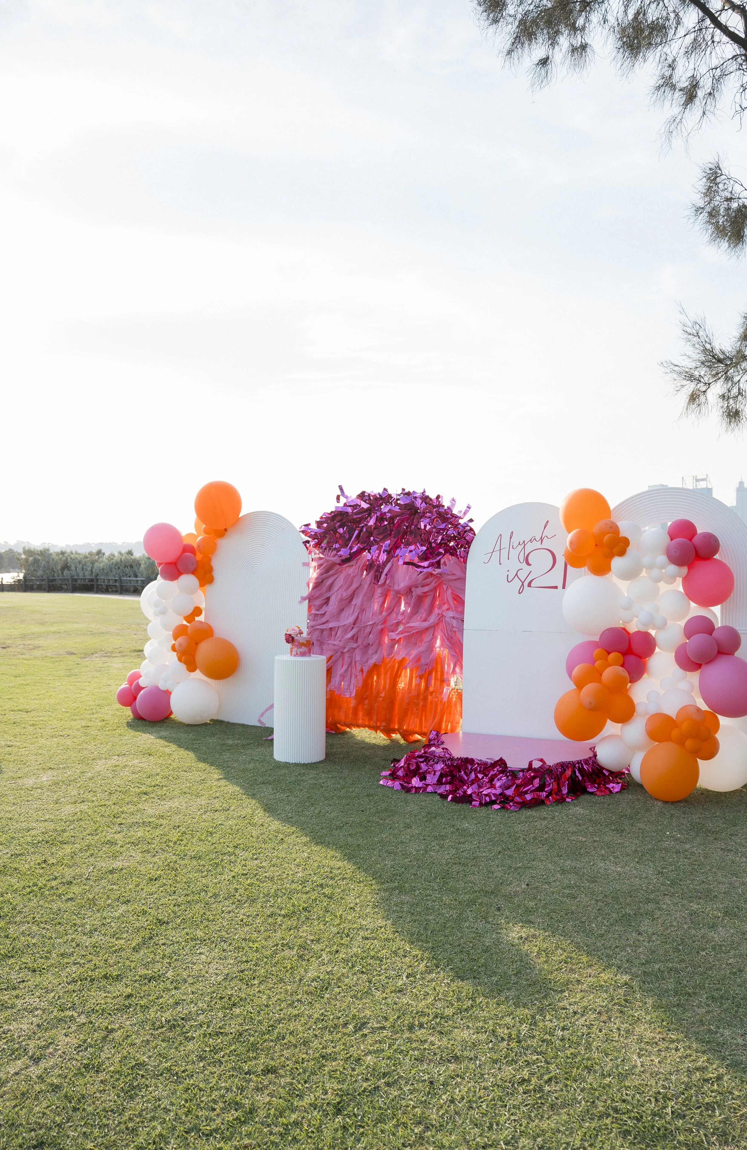 Outdoor celebration setup with pink, orange, white balloons, pink fringe backdrop, a white sign with pink writing that says 'Aiyah 12', and a grassy field with trees and a fence in the background.