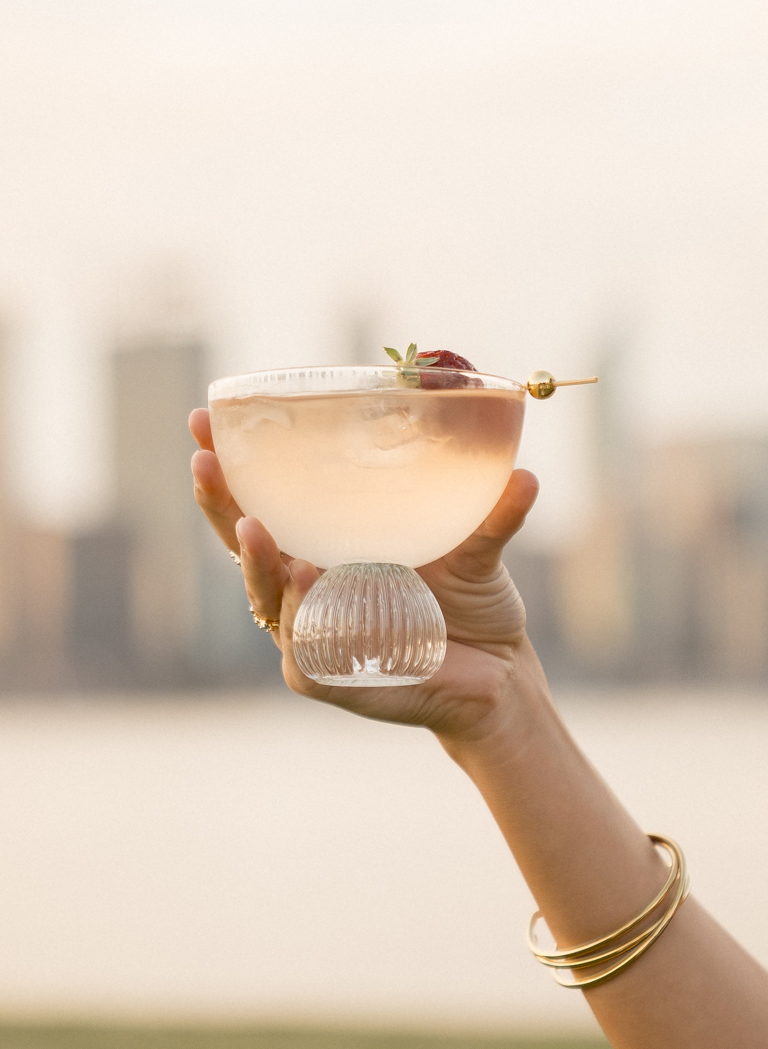 A person's hand holding a glass bowl filled with a pink beverage garnished with a strawberry and mint leaf, with a skewer and rings on the wrist.