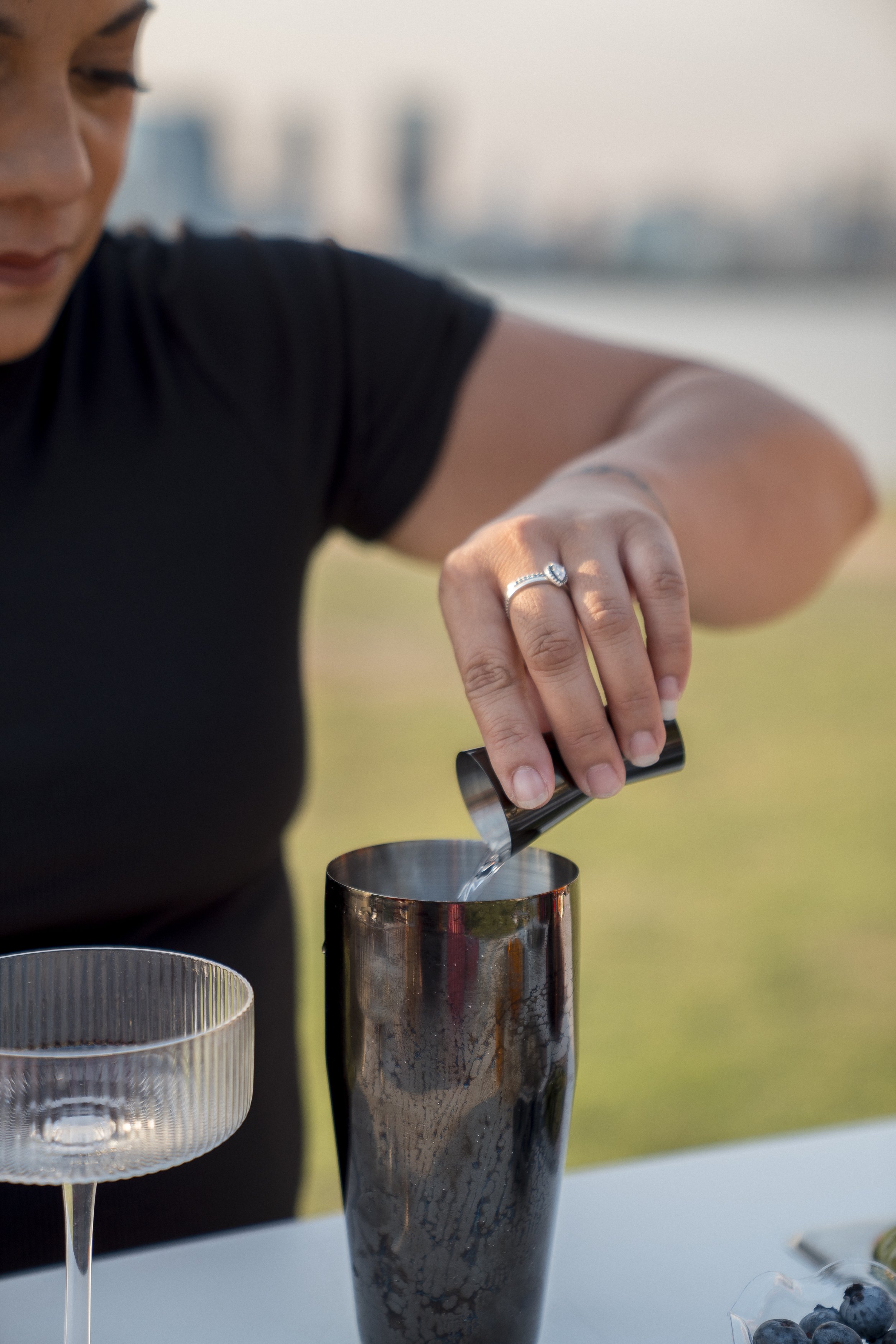 Person pouring a drink from a cocktail shaker into a tall glass, with another empty glass nearby and a bowl of blueberries on the table.