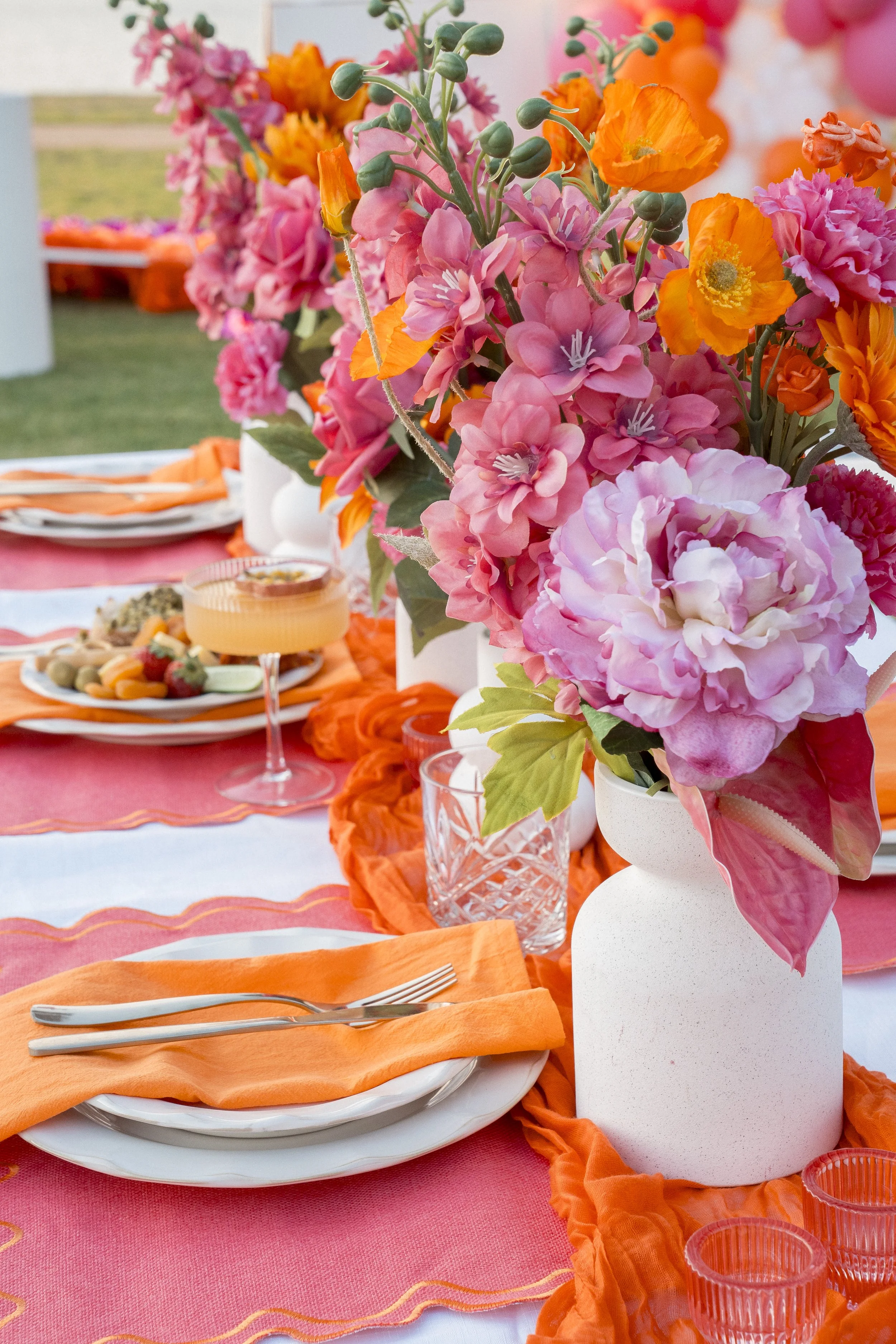 Vase with pink peonies and orange poppies on a table with pink and orange tableware at an outdoor event.