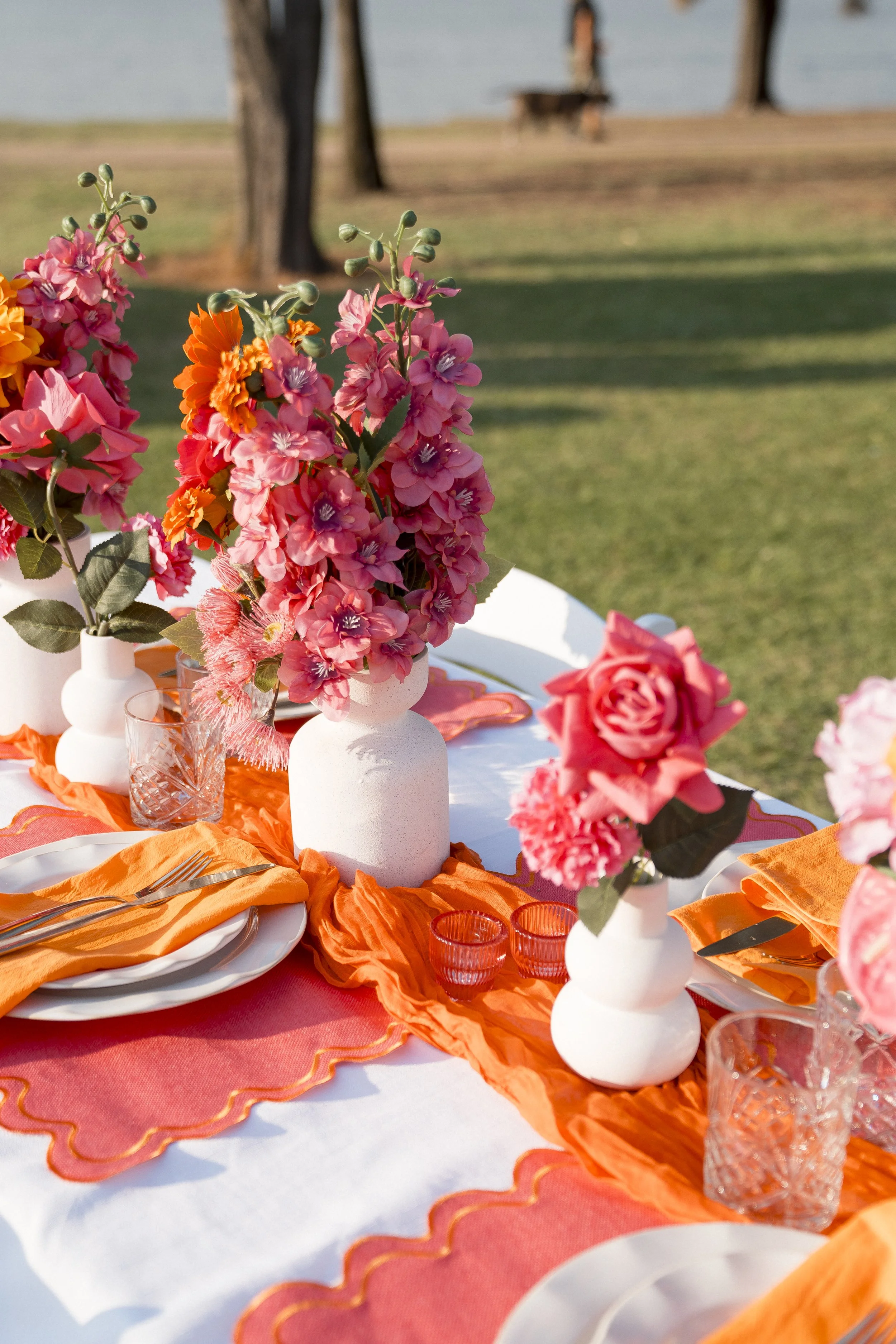 Table decorated with pink and orange flowers in white vases, orange and pink tablecloths, place settings, and glasses outdoors by the water.