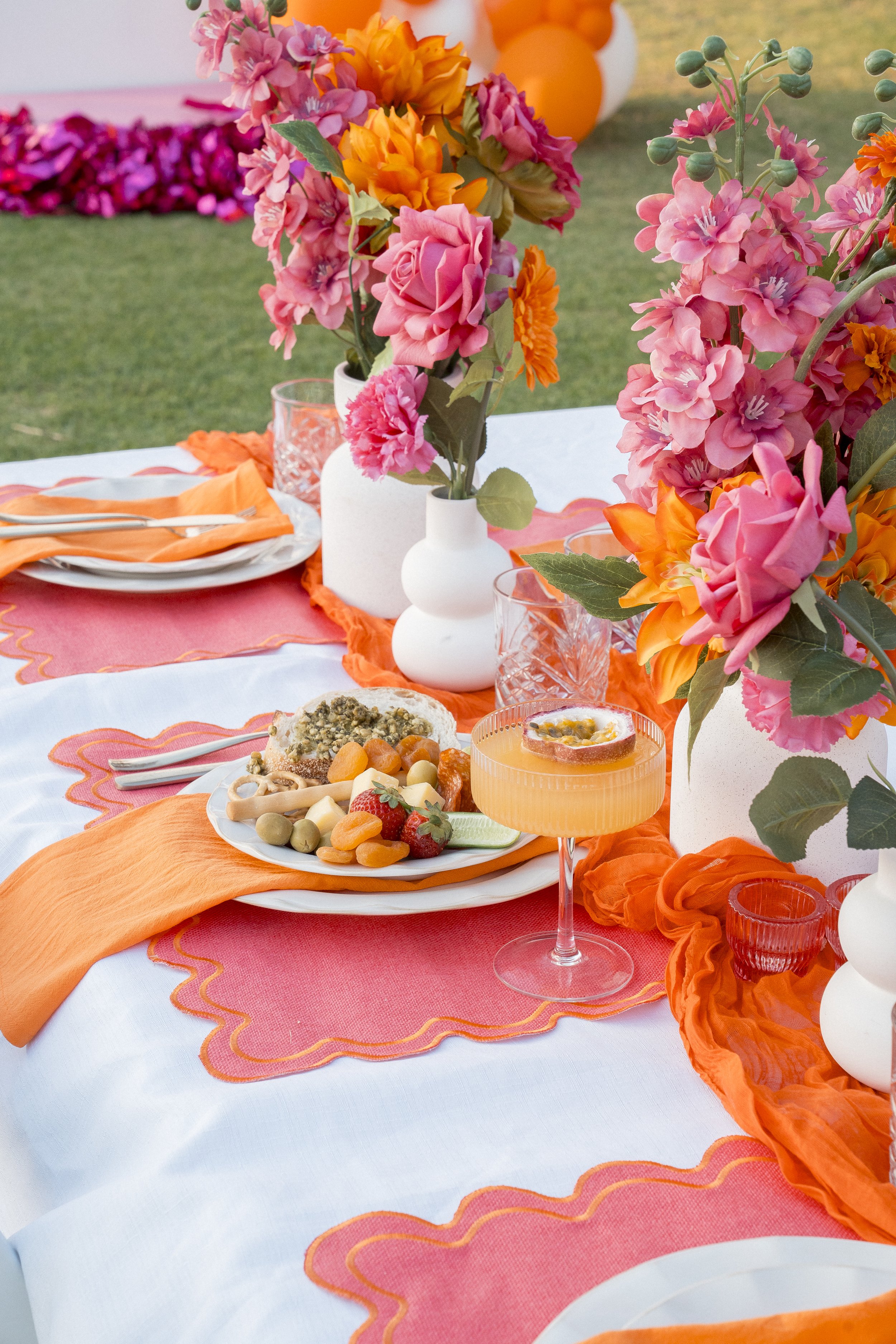A decorated outdoor table with pink, orange, and peach floral centerpieces in white vases, set with orange cloth napkins, glassware, and plates of food, with a grassy background.