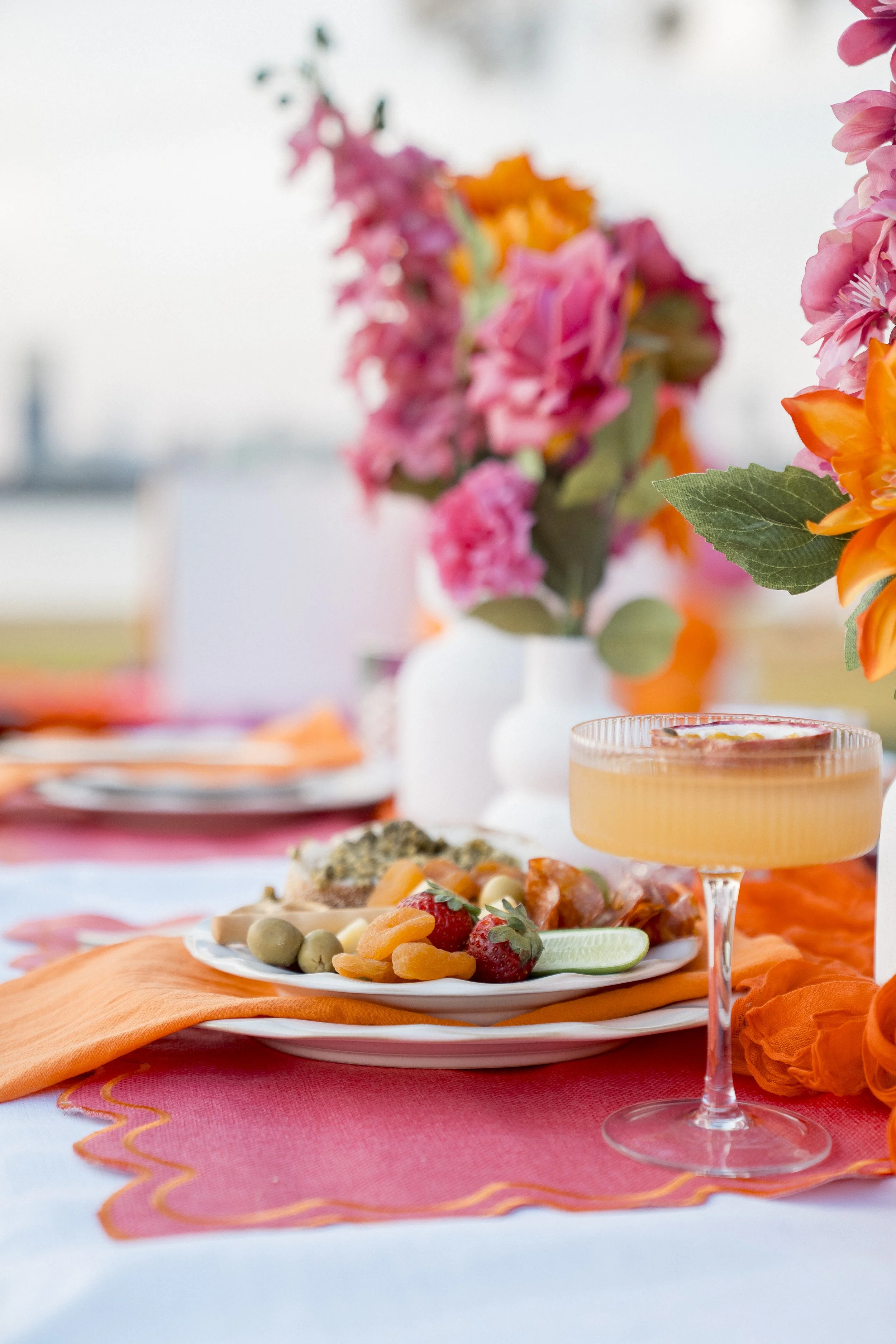 Close-up of a table with a white plate of fruit including strawberries, green olives, and sliced cucumber, with an orange cloth underneath. There is a pink and orange flowers centerpiece and a pink cocktail in a tall glass on the table.