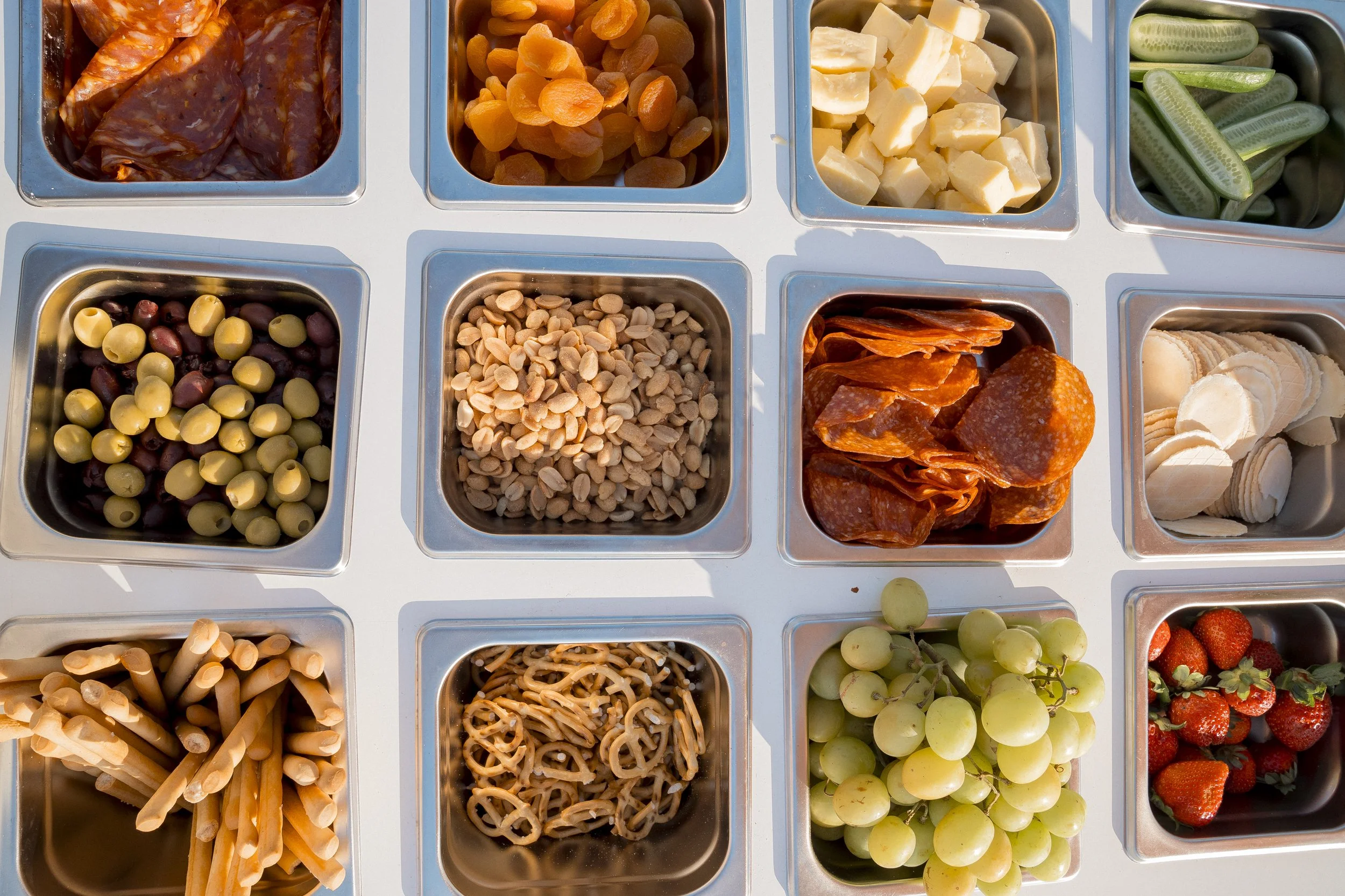 A selection of toppings at an ice cream or frozen yogurt bar, including chocolate chips, dried apricots, diced cheese, cucumber slices, mixed beans, chopped almonds, pretzels, sausage slices, white grapes, strawberries, and other fruits.