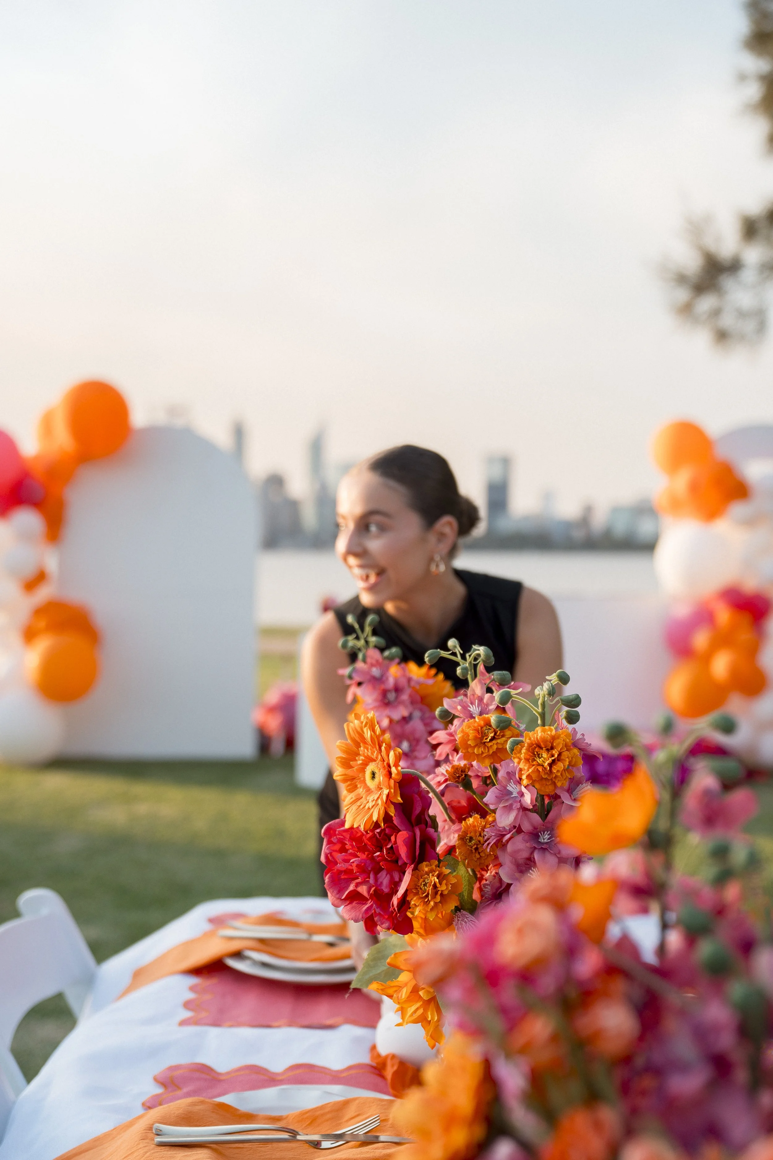A woman with dark hair tied back, smiling, at a beautifully decorated outdoor event table with a city skyline in the background. The table features vibrant orange and pink flowers, white tablecloths, and orange accents.