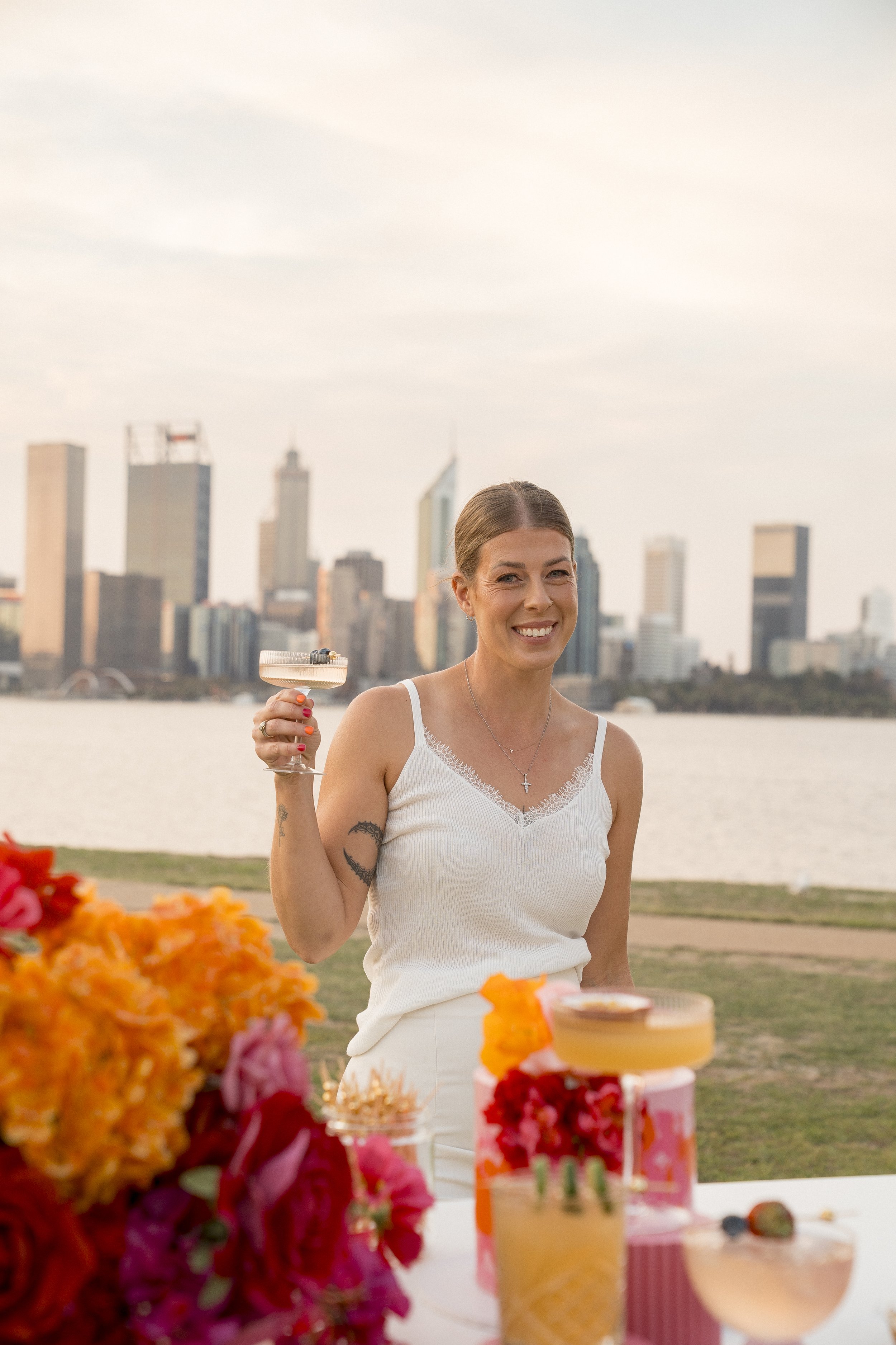 A woman smiling and holding a cocktail glass at an outdoor celebration, with a city skyline in the background and a table of colorful desserts in the foreground.