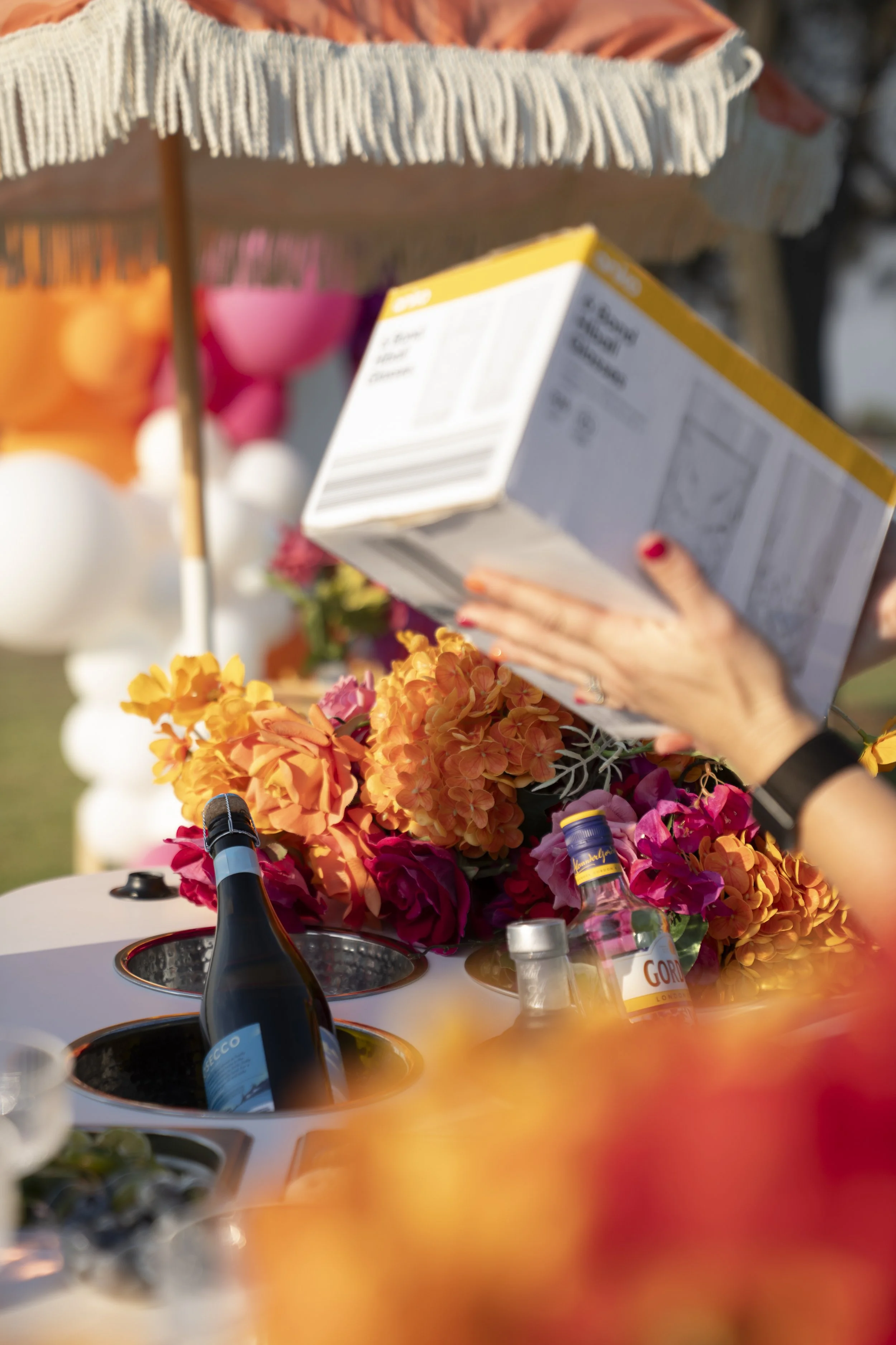 A person holding a menu at a festive outdoor event with colorful flowers and bottles on a table, under a cream-colored fringed umbrella.