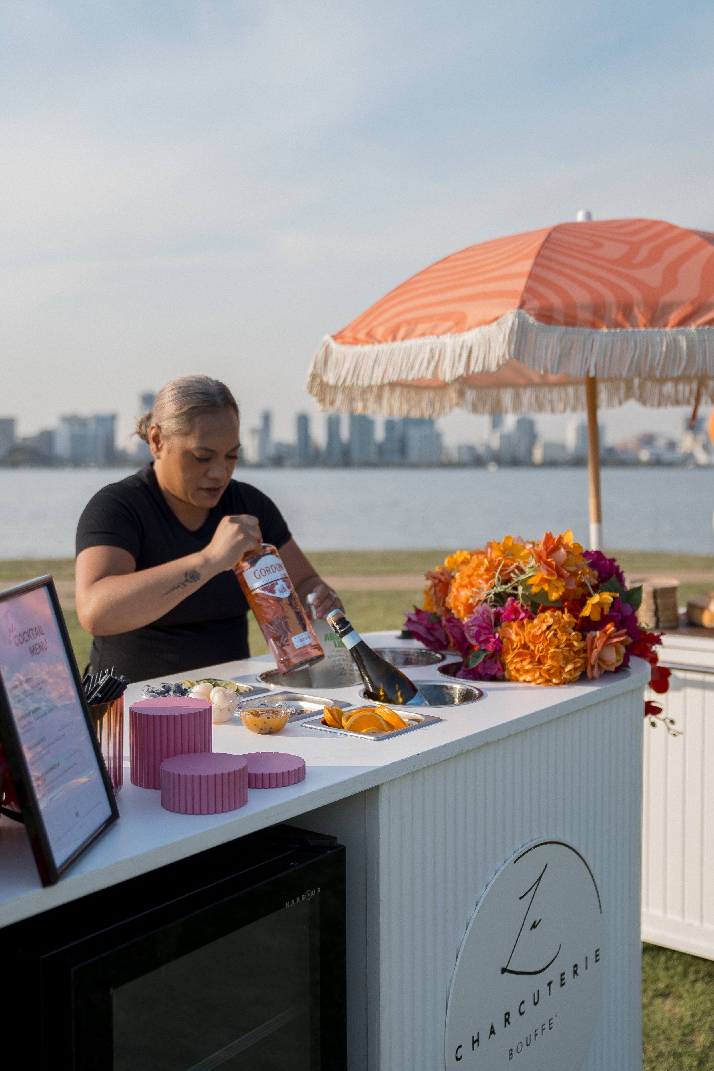 A woman preparing drinks at an outdoor juice bar with a scenic city skyline in the background. The juice bar is decorated with colorful flowers and is shaded by a orange and white fringed umbrella.
