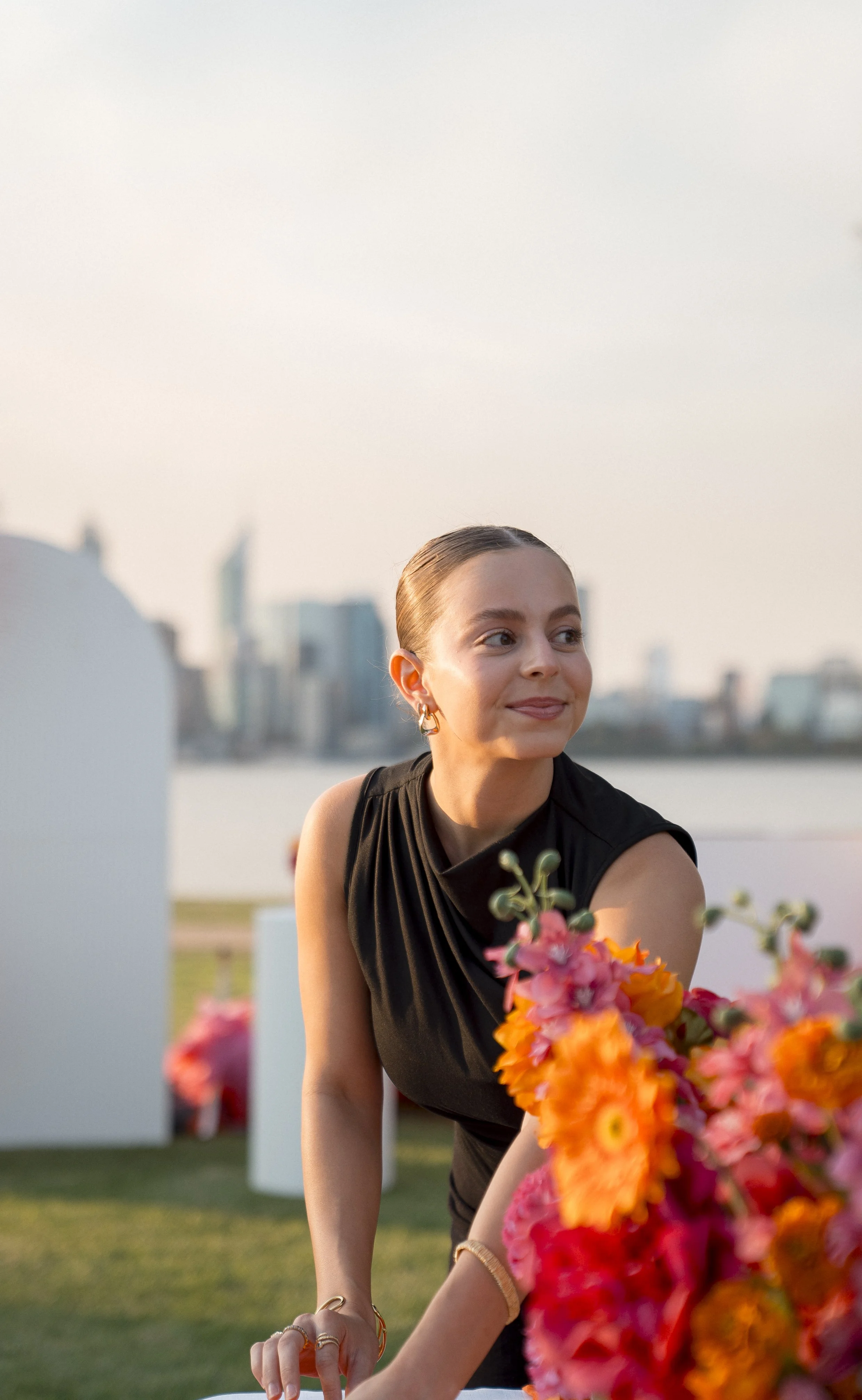 A young woman with slicked-back hair and gold earrings, wearing a sleeveless black dress, arranging a colorful bouquet of flowers outdoors against a city skyline backdrop during sunset.