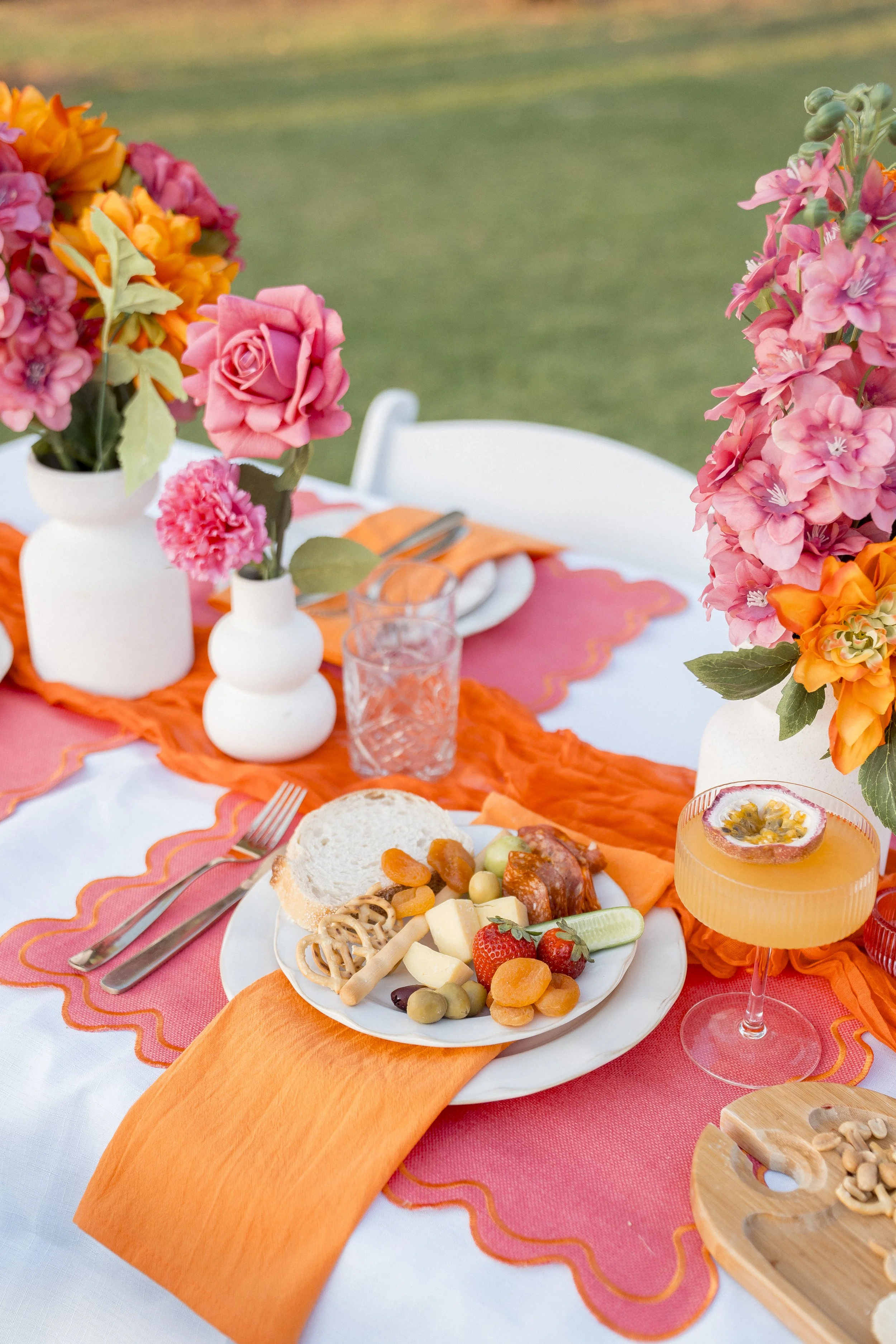 A table set for an outdoor meal with colorful flower arrangements in white vases, a plate of assorted fruits, bread, and cheese, orange napkins, and pink and orange tablecloths, with a grassy background.